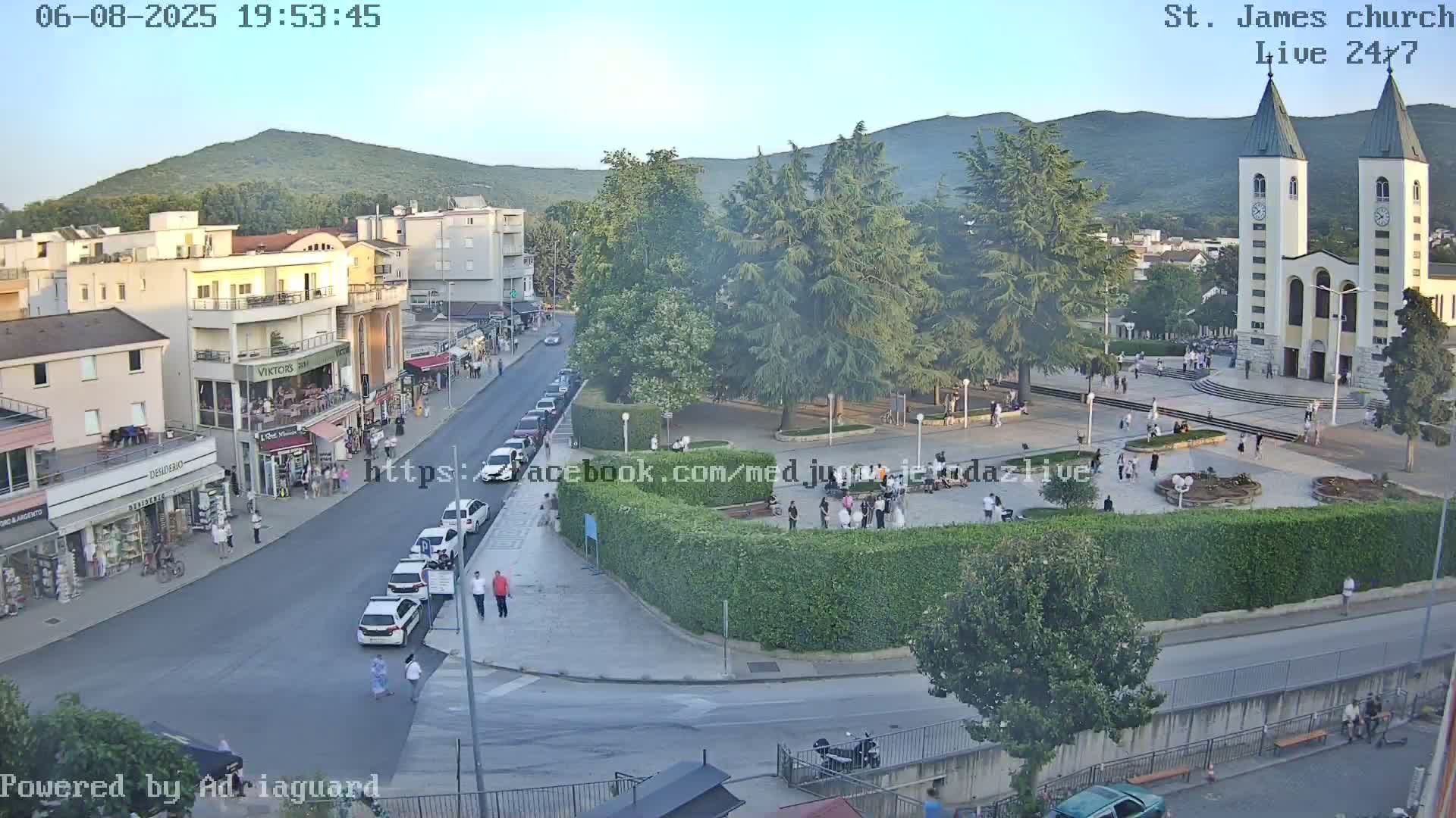 An aerial view of a town square at dusk, showing a church, parked cars, pedestrians, and lush greenery under a clear sky.