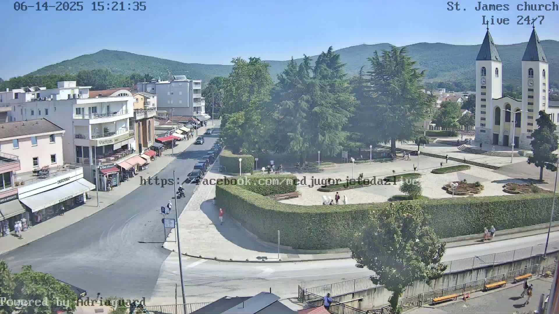 A sunny day view of a town square with a church, buildings, and a few people, surrounded by greenery and mountains in the distance.