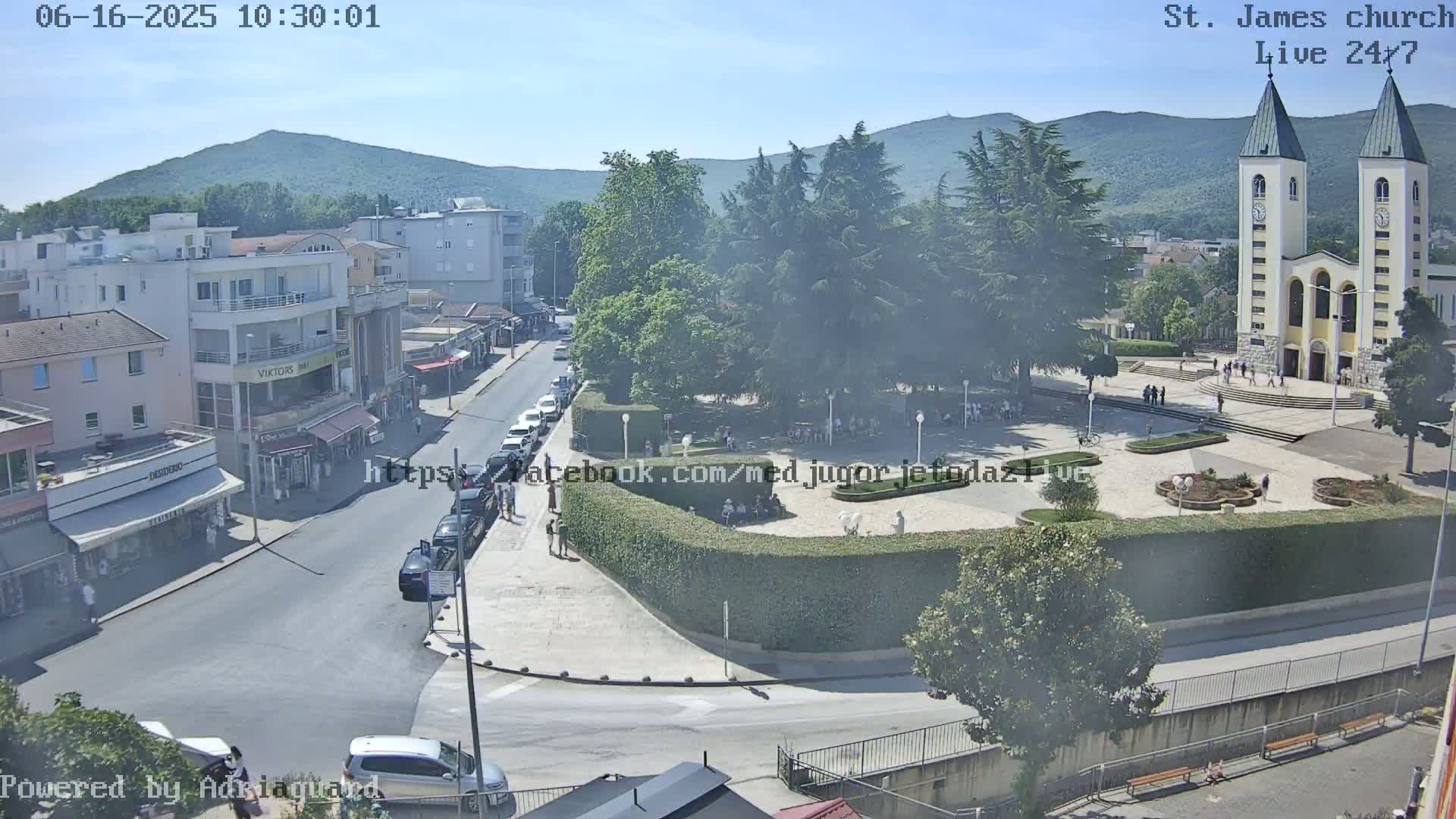 A high-angle, long shot of a town square on a sunny day, centered on a large church with twin spires, surrounded by buildings, parked cars, and people.
