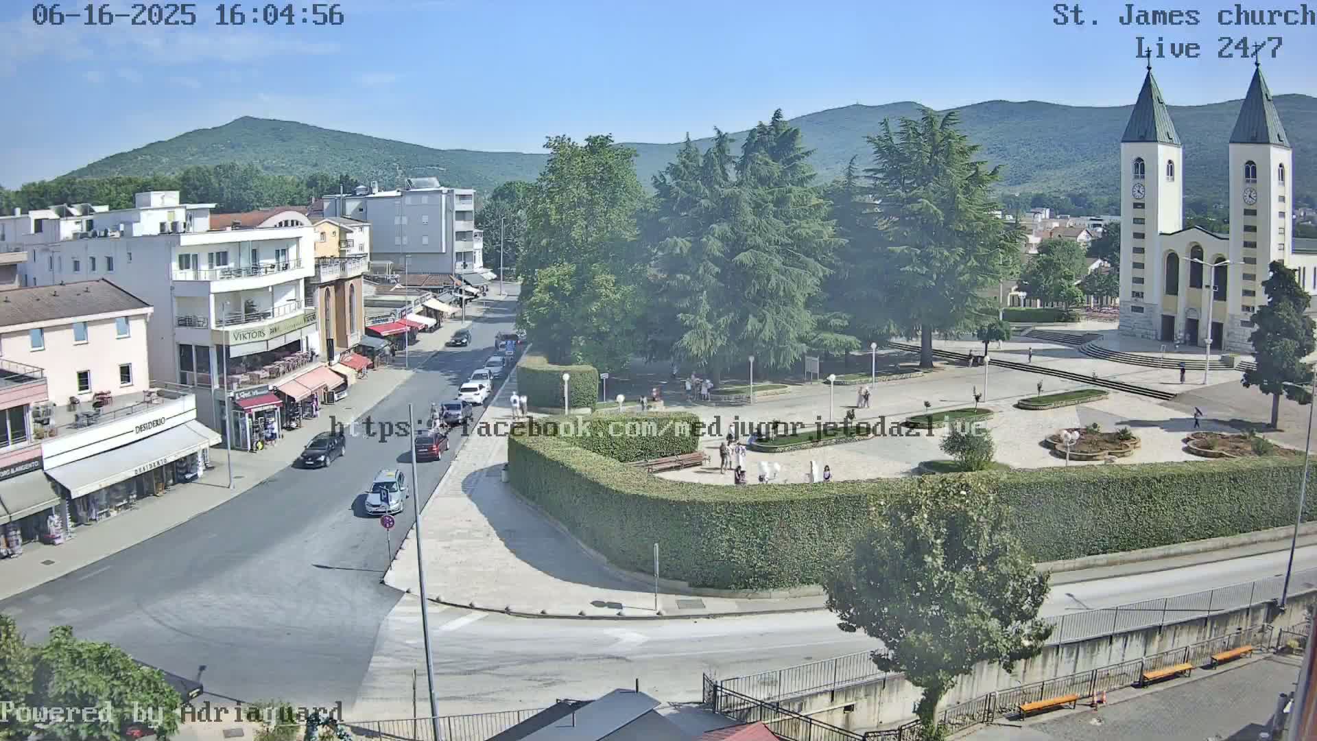 A sunny view of a town square with a church, buildings, cars, and people,  surrounded by lush greenery and mountains in the background.