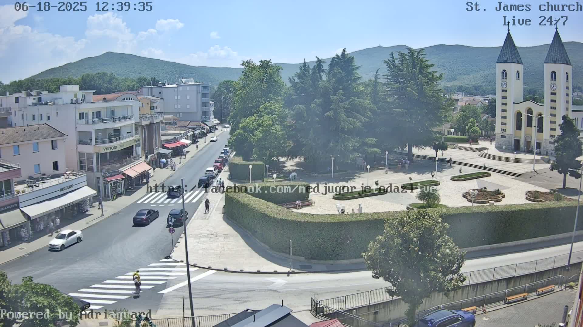 A sunny day reveals a town square with a church, surrounded by buildings, trees, and cars, with a few pedestrians visible.