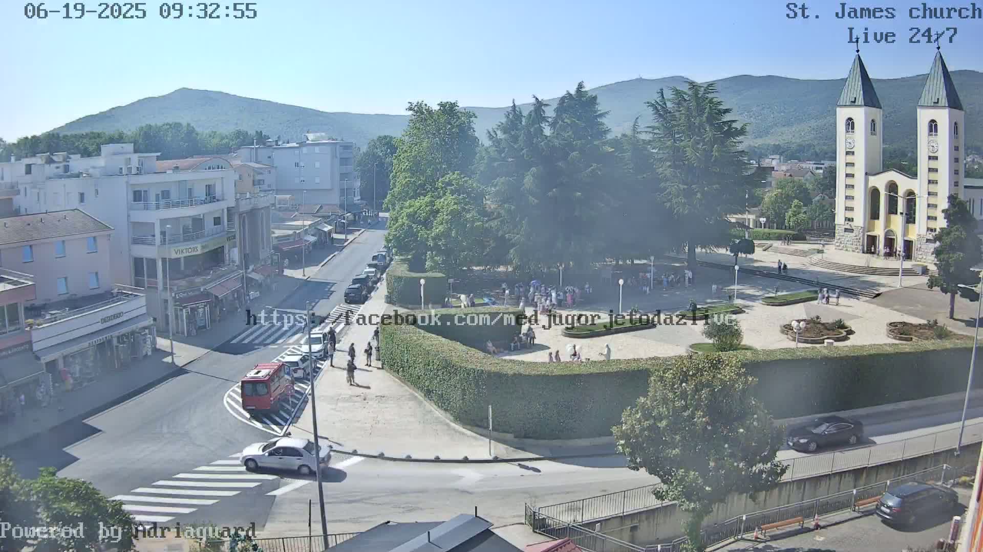 A sunny day reveals a town square with a church, several buildings, cars, and pedestrians, surrounded by lush greenery and mountains in the distance.