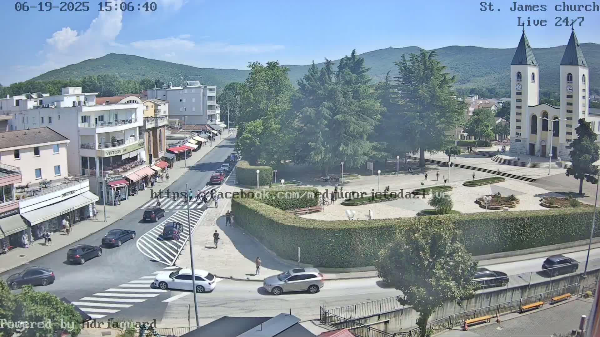A sunny day shows a town square with a church, cars on roads, pedestrians, and green spaces, all surrounded by buildings and hills.