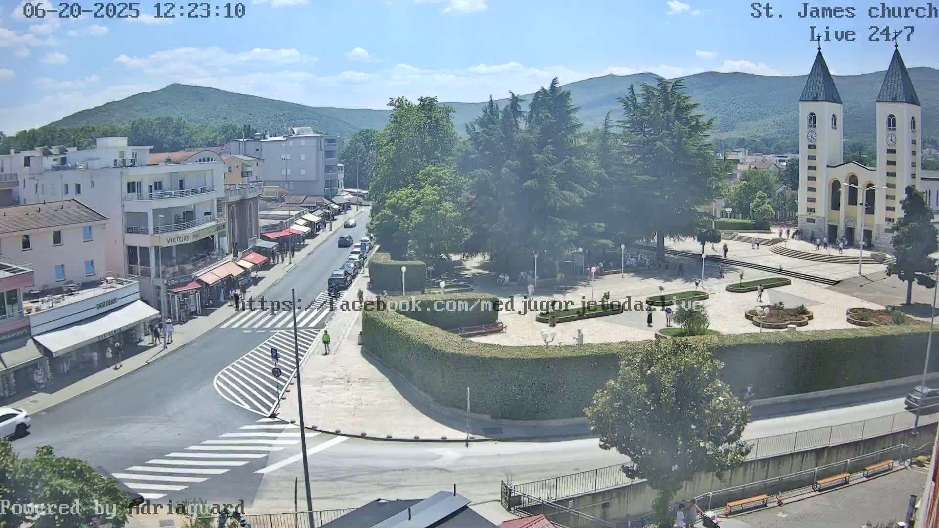 A sunny day shows a town square with a church, surrounding buildings, and a few people; mountains are visible in the background.