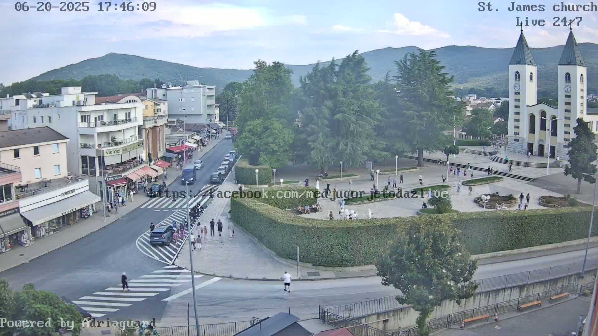 A high-angle, long shot shows a town square with a church, buildings, people walking around, cars parked along the street, and lush green trees under a partly cloudy sky.
