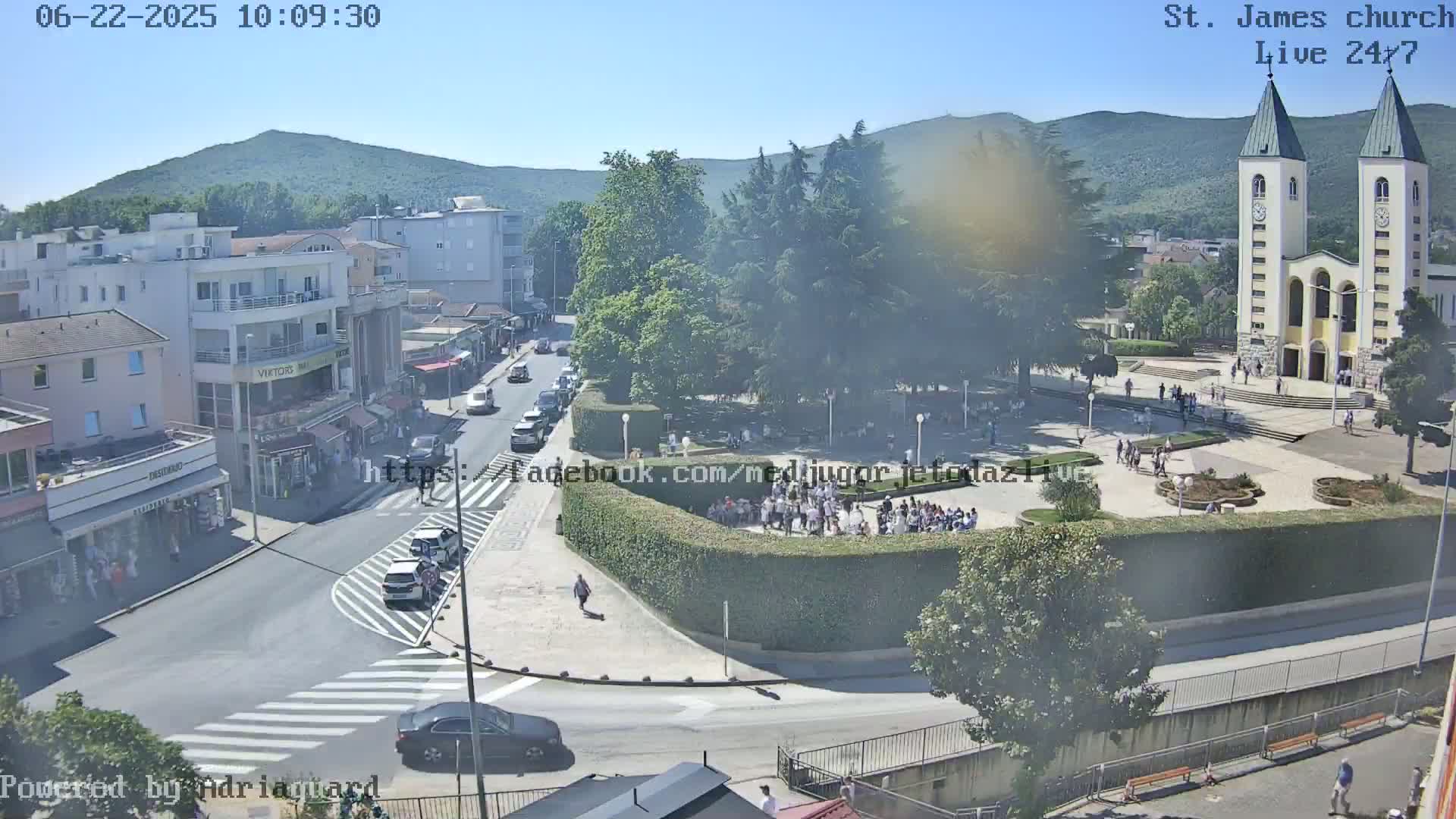 A sunny day reveals a town square with a large church, surrounded by buildings, cars, and people; a hill is visible in the background.