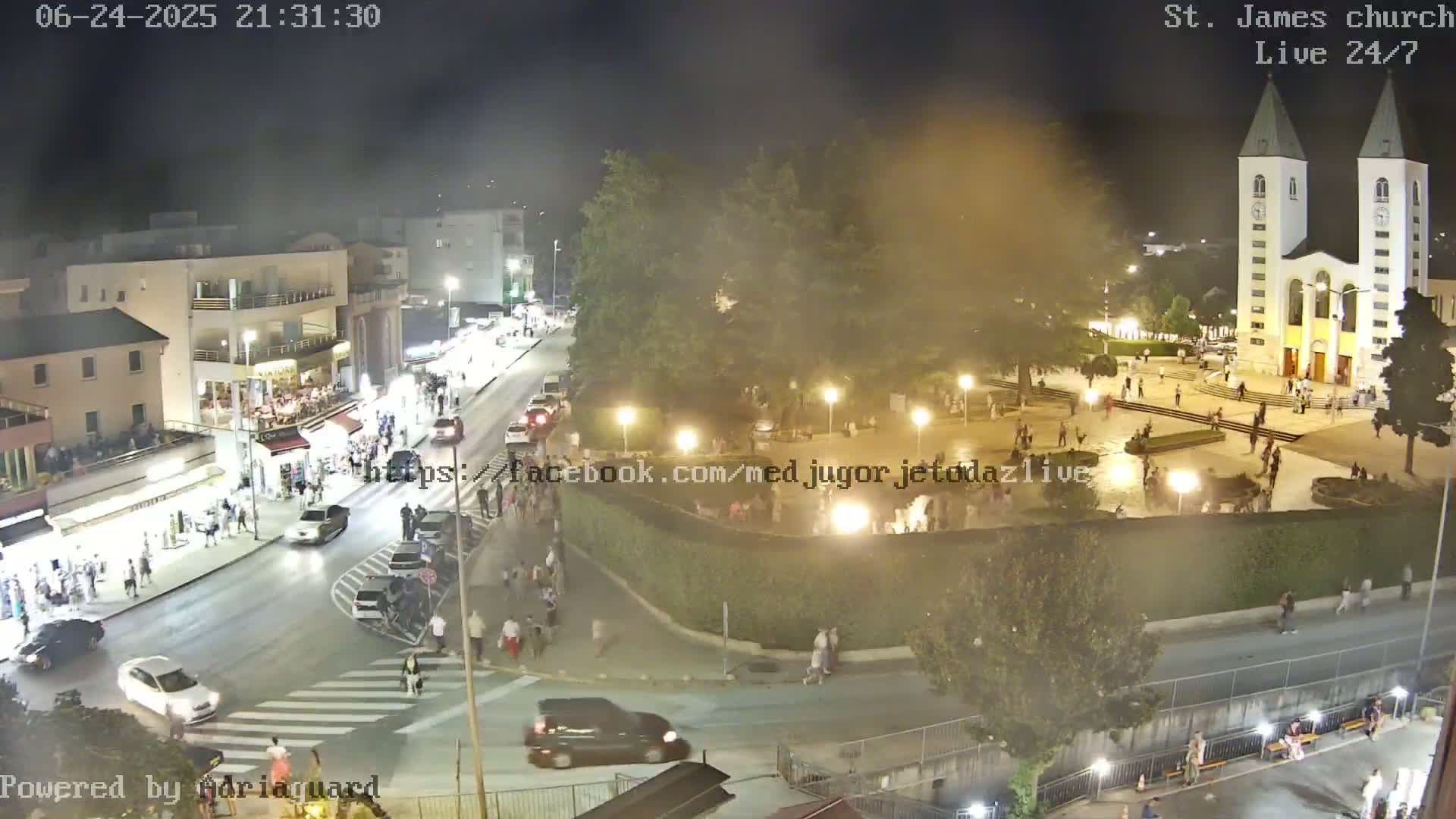 A nighttime aerial view of a town square with a church, many people, and cars on the streets under hazy conditions.