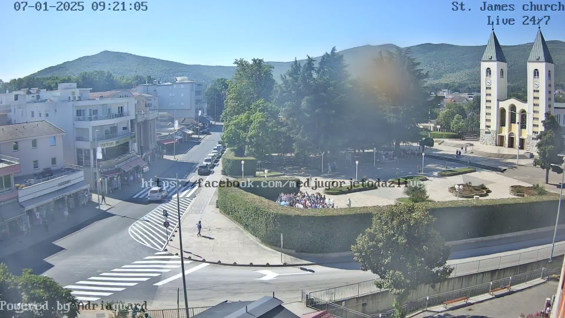 A high-angle view shows a town square on a clear day with a large church in the background, surrounded by buildings and green spaces.