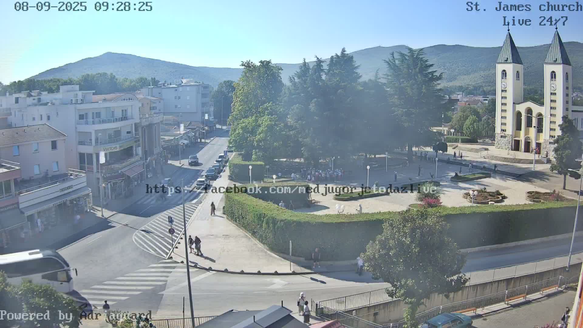 A high-angle, long shot on a sunny day shows a town square with a large church in the background, surrounded by buildings, cars, and pedestrians.
