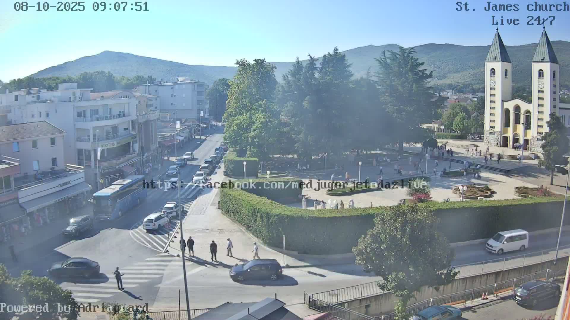 A high-angle, long shot on a sunny day shows a town square with a church, pedestrians, vehicles, and buildings, all surrounded by greenery and mountains in the distance.