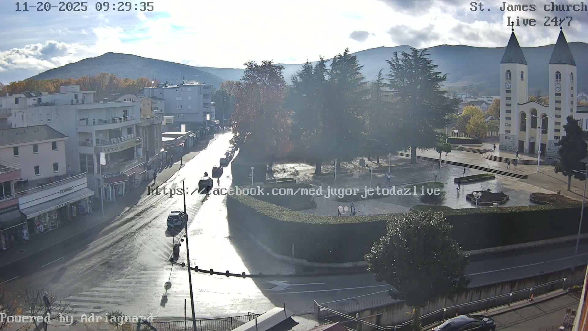 An overhead view captures a vibrant town square and street, featuring a large twin-spired church, surrounding buildings, and distant mountains, all under a bright, partly cloudy sky that casts reflections on the wet ground from recent rain, with a few pedestrians and cars moving about.
