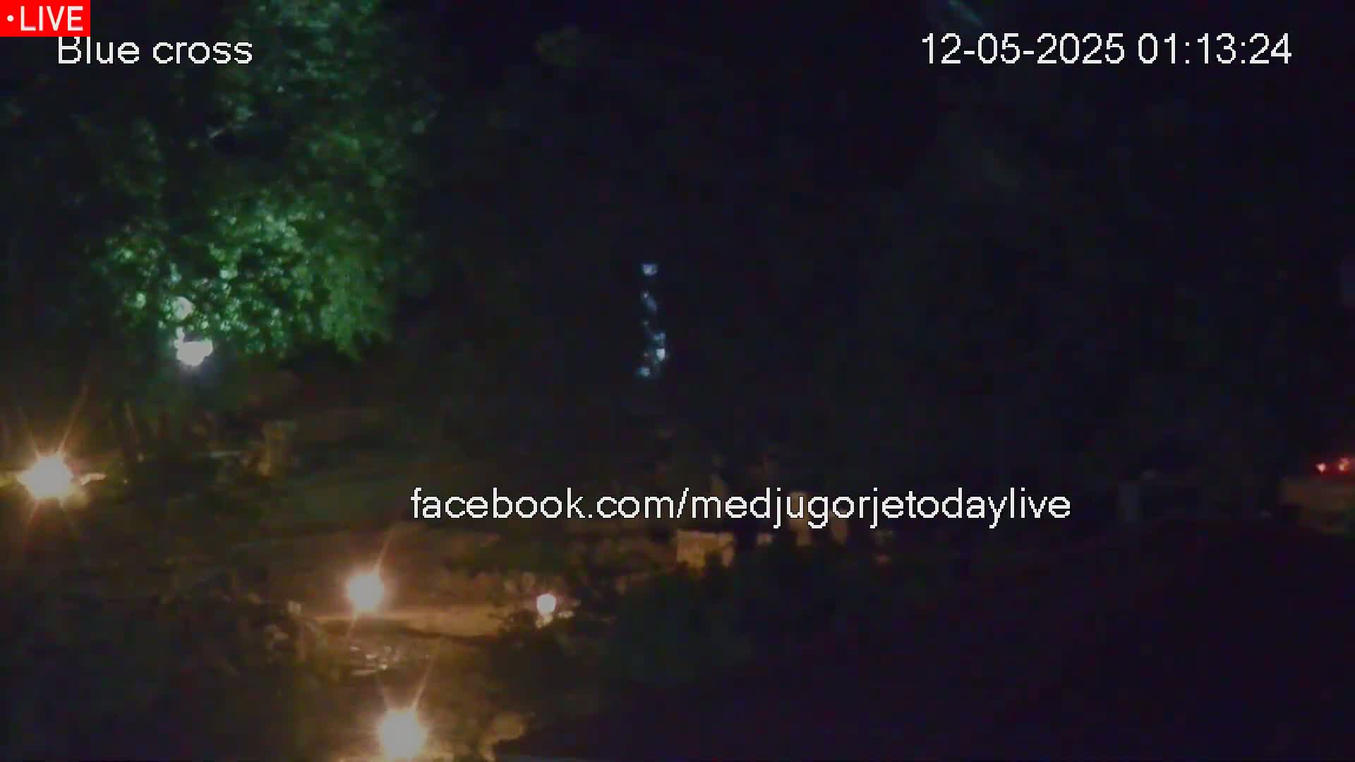 At night, several small, flickering lights illuminate a dark area with trees and a body of water.