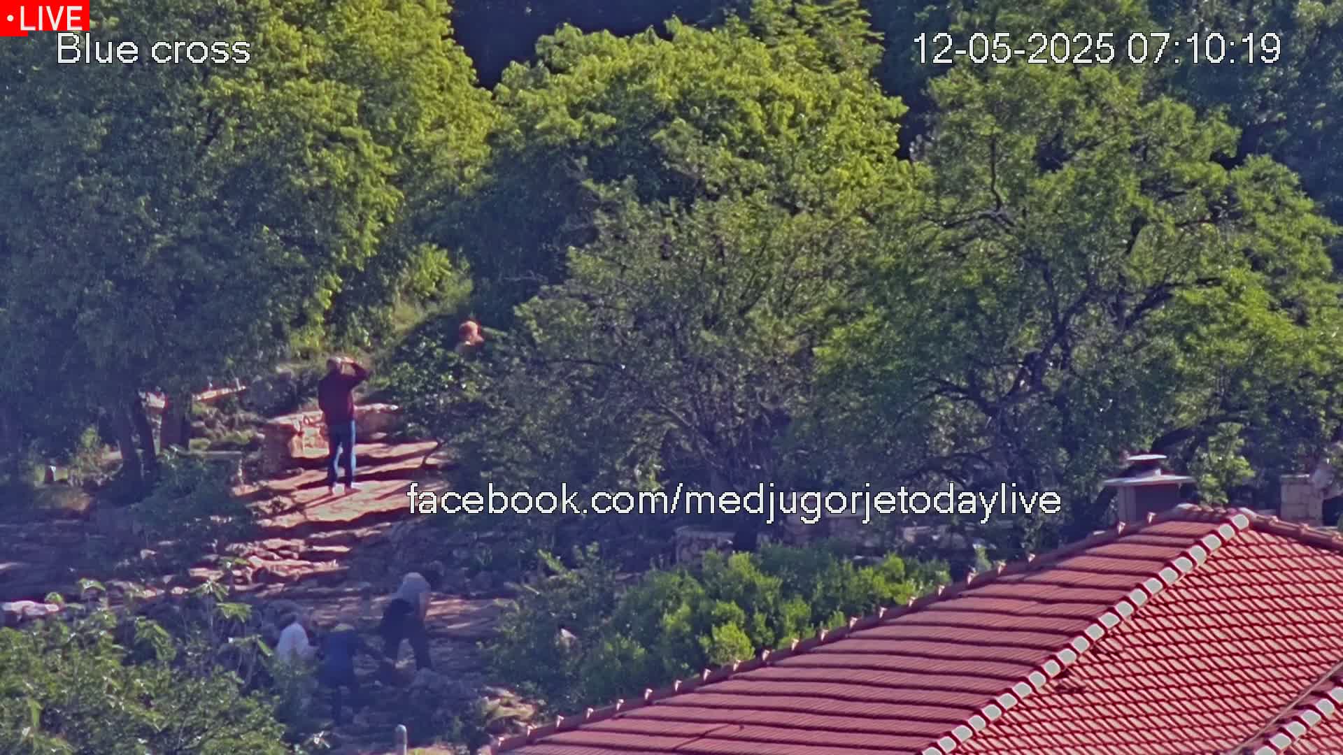 A sunny outdoor scene shows several people standing on a rocky path amongst lush green trees and a red-tiled roof.