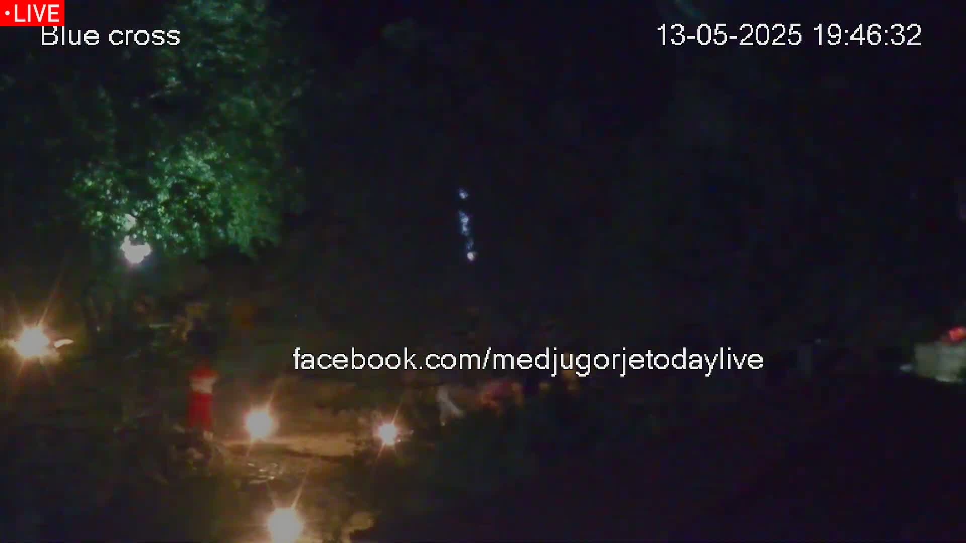 At night, several small lights illuminate a dark scene with people gathered near a tree under a clear sky.