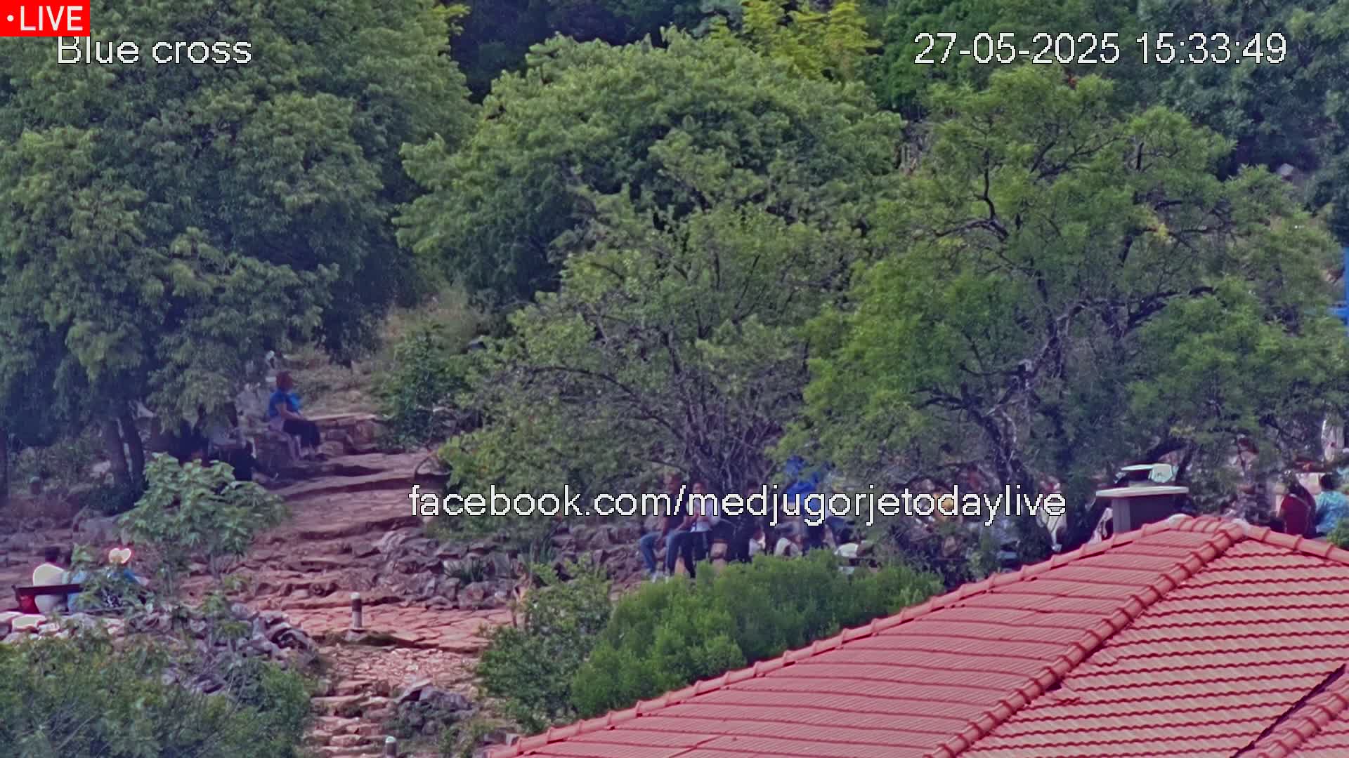 A rocky, tree-filled area with a group of people gathered near a red-tiled roof, under a partly cloudy sky.