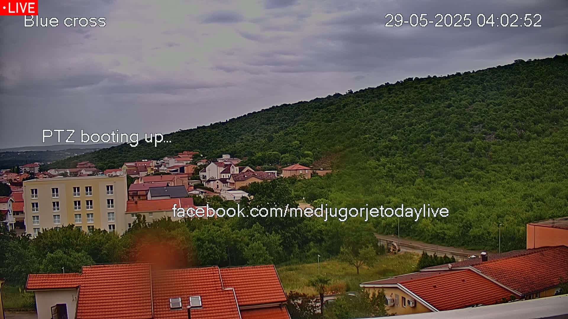 An overcast day shows a view of a town nestled at the base of a lush green hill, with red-tiled roofs dominating the buildings.
