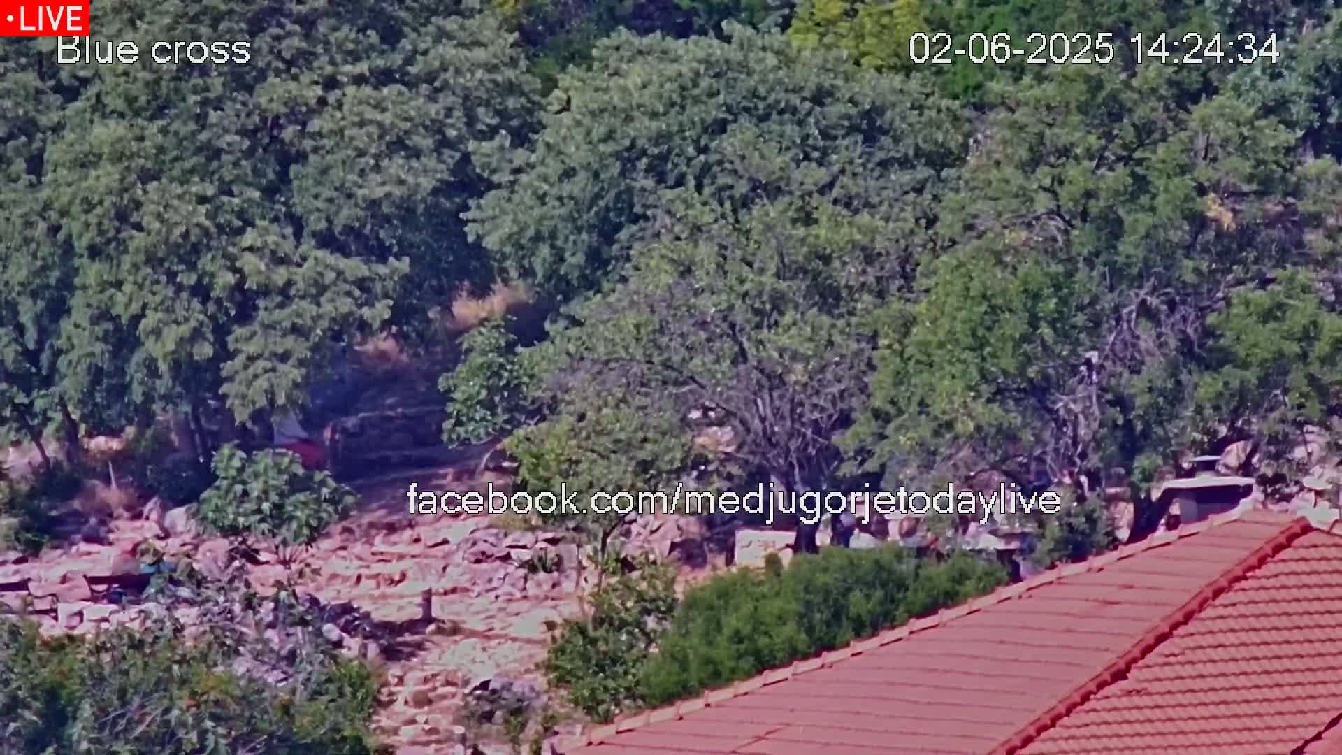 A rocky, wooded area with a red-tiled roof in the bottom right corner is visible on a sunny day.
