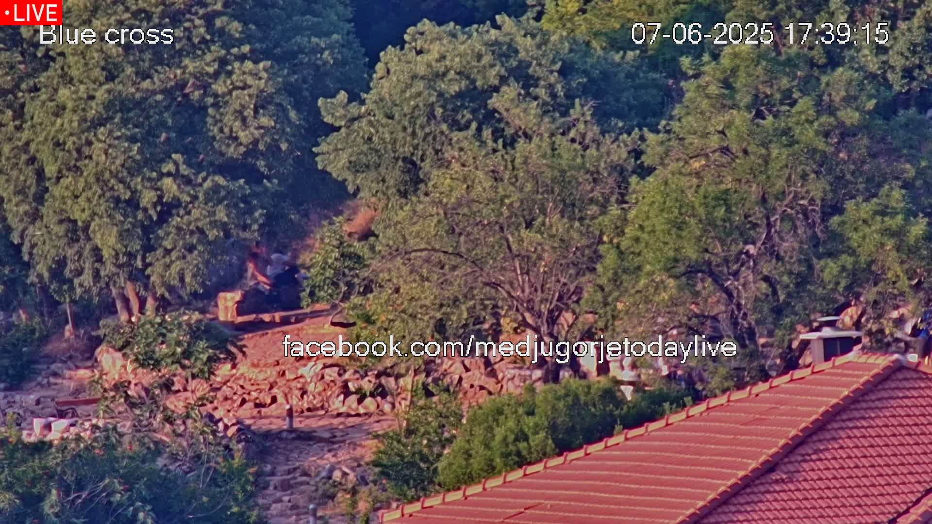 A high-angle, long shot shows a rocky, wooded area with a few people near a pile of rocks, and a reddish-brown roof in the foreground under a sunny sky.