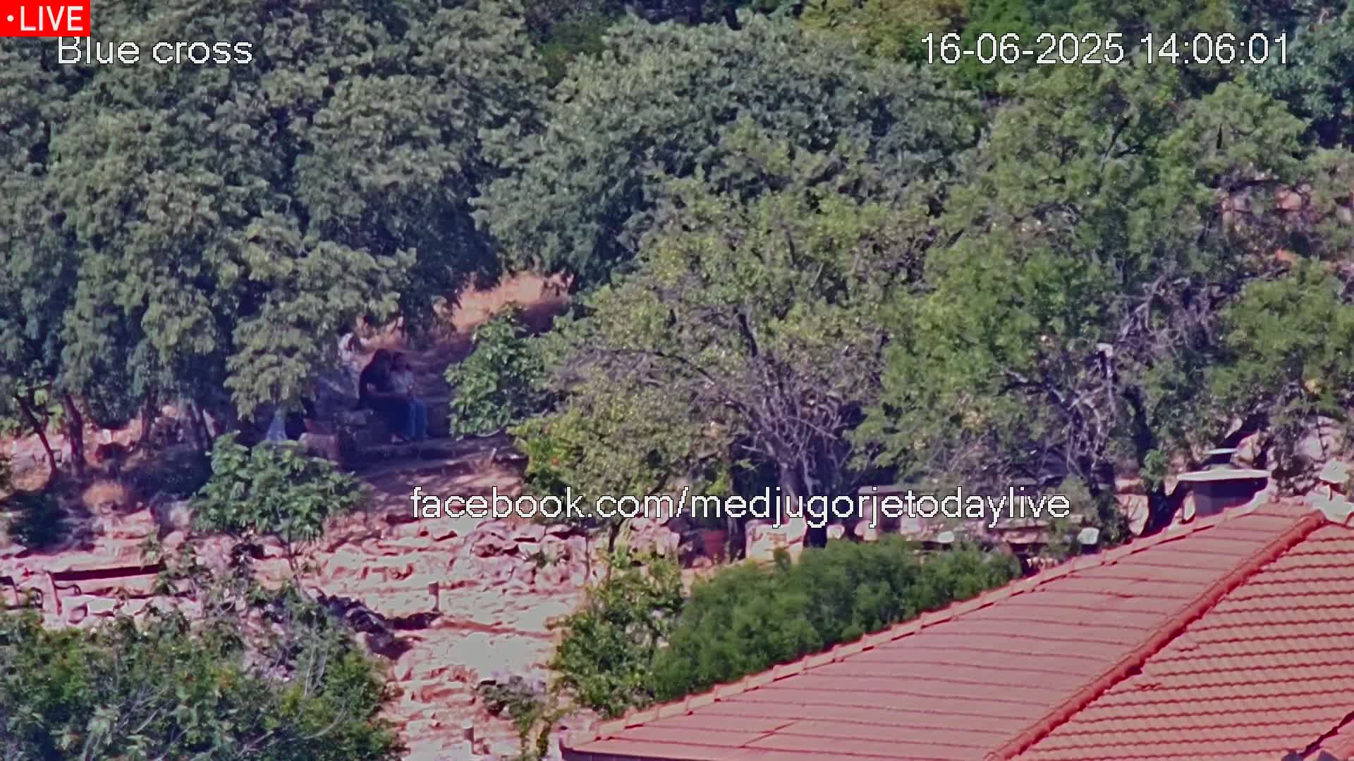 A high-angle view shows a rocky, wooded area with several people sitting beneath some trees, and a red-tiled roof in the foreground, under sunny conditions.