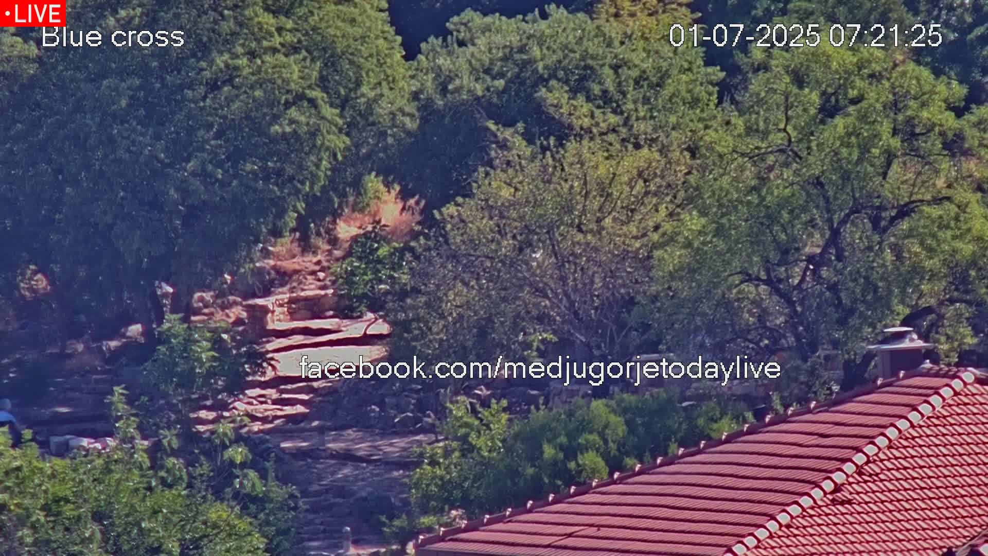A rocky path winds through a lush, green landscape under a sunny sky, partially visible behind a red-tiled roof.