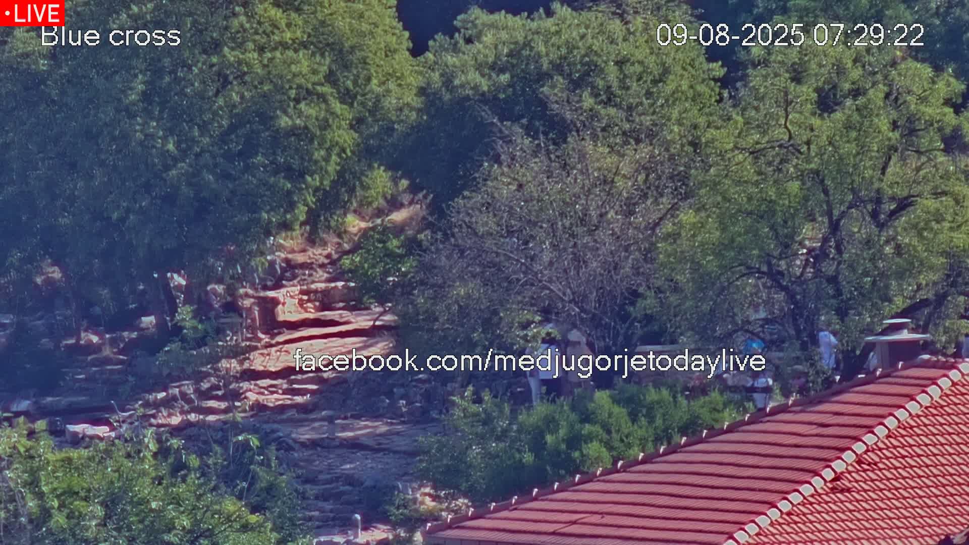 A rocky hillside covered in green trees is partially visible behind a red-tiled roof, under a sunny sky.
