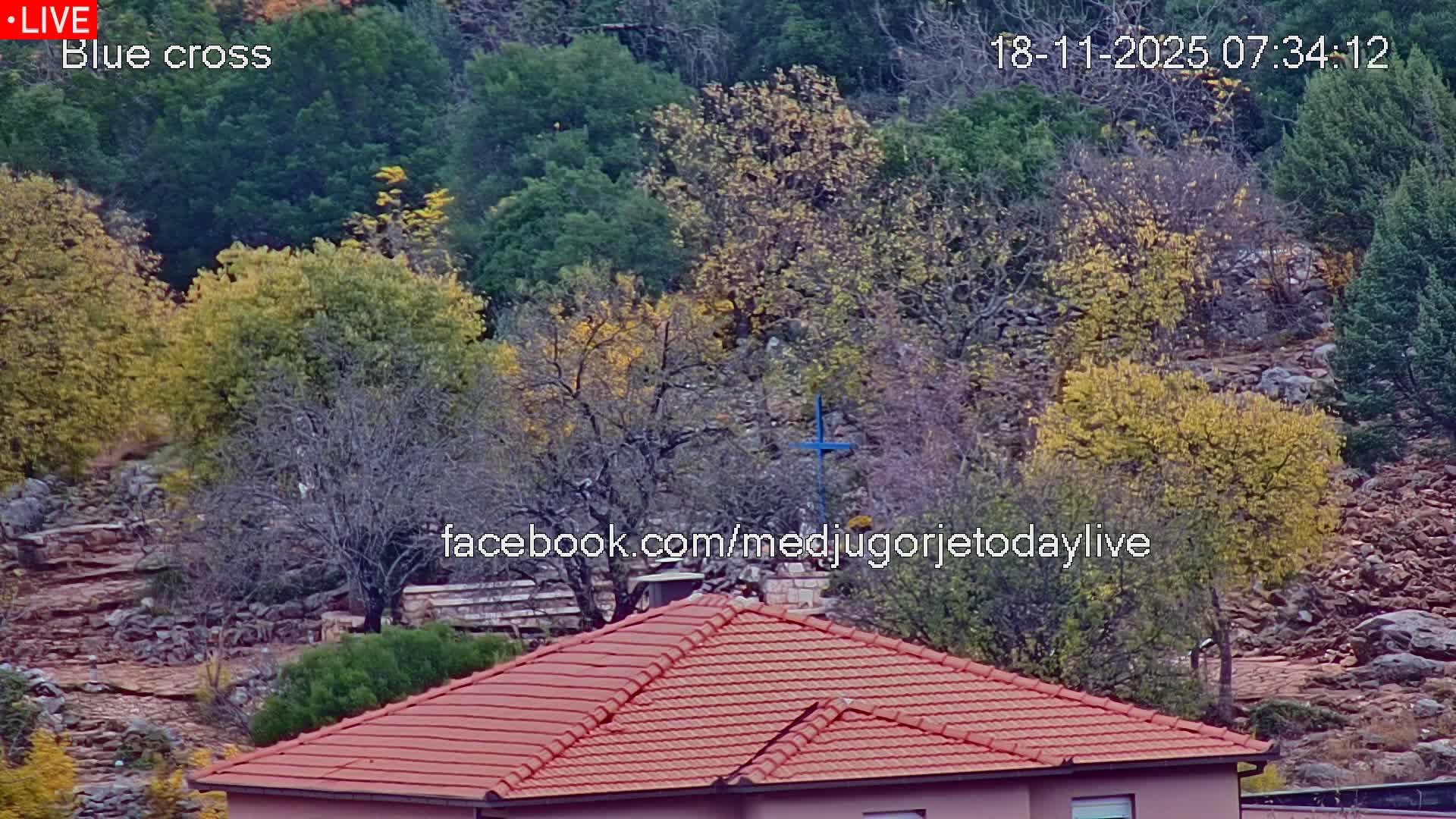 A rocky hillside covered in green trees is partially visible behind a red-tiled roof, under a sunny sky.