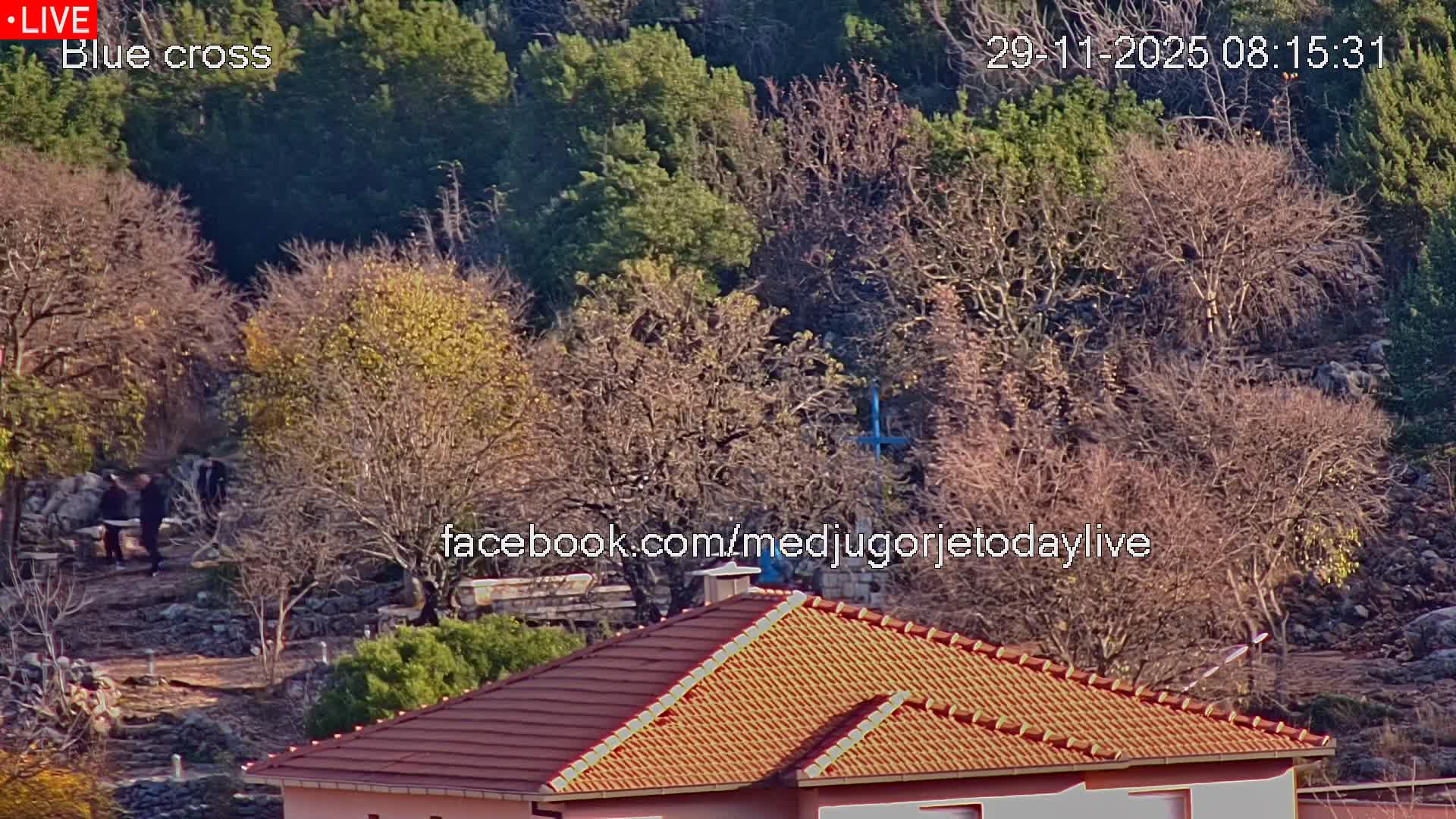 The image displays a sunny, clear outdoor scene featuring a terracotta-tiled roof in the foreground, a path winding through a landscape of mostly bare deciduous trees and evergreens, with a blue cross and several people visible in the middle ground.