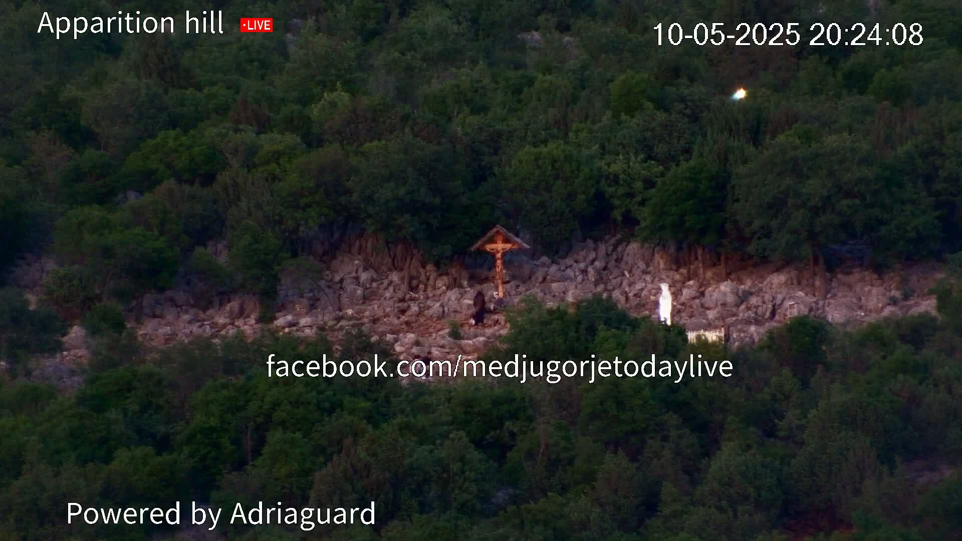 A rocky hillside covered in dark green trees, with a crucifix and a statue visible near its center, under what appears to be a twilight sky.