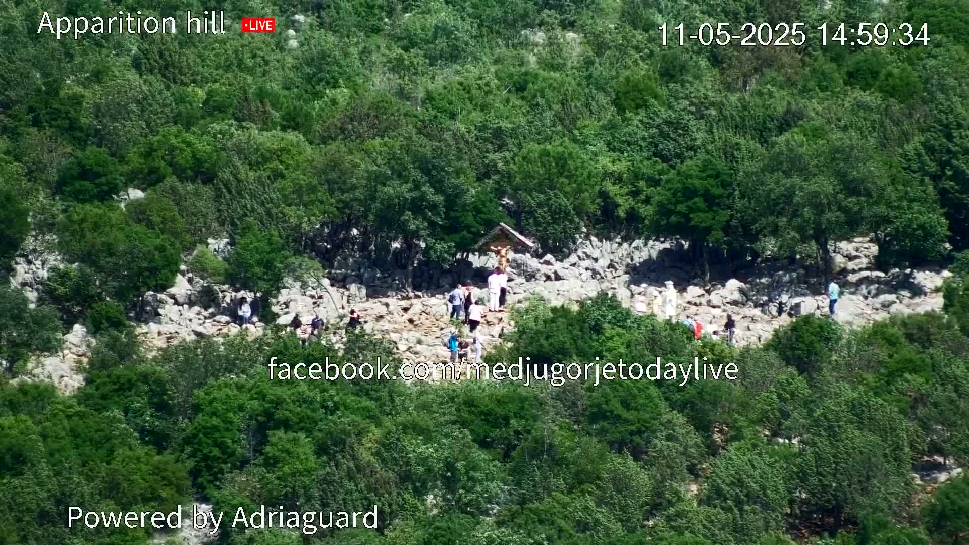 A rocky hillside covered in lush green vegetation shows a group of people gathered near a small religious structure on a sunny day.