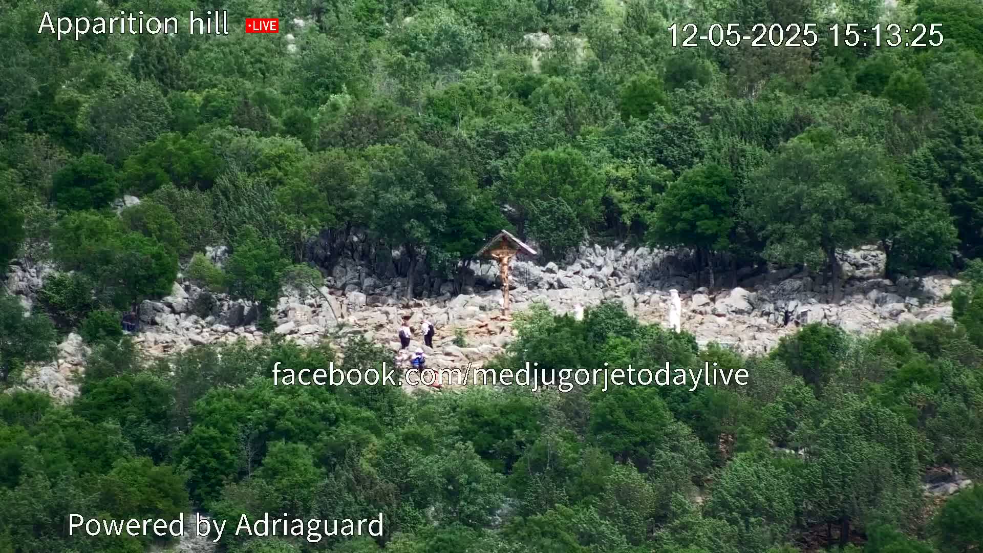 A rocky hillside covered in green vegetation shows a few people near a crucifix.