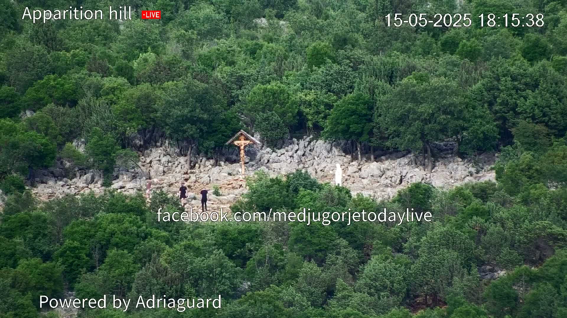 A rocky hillside covered in lush green vegetation features a crucifix and a statue, with several people standing near them, under a clear sky.