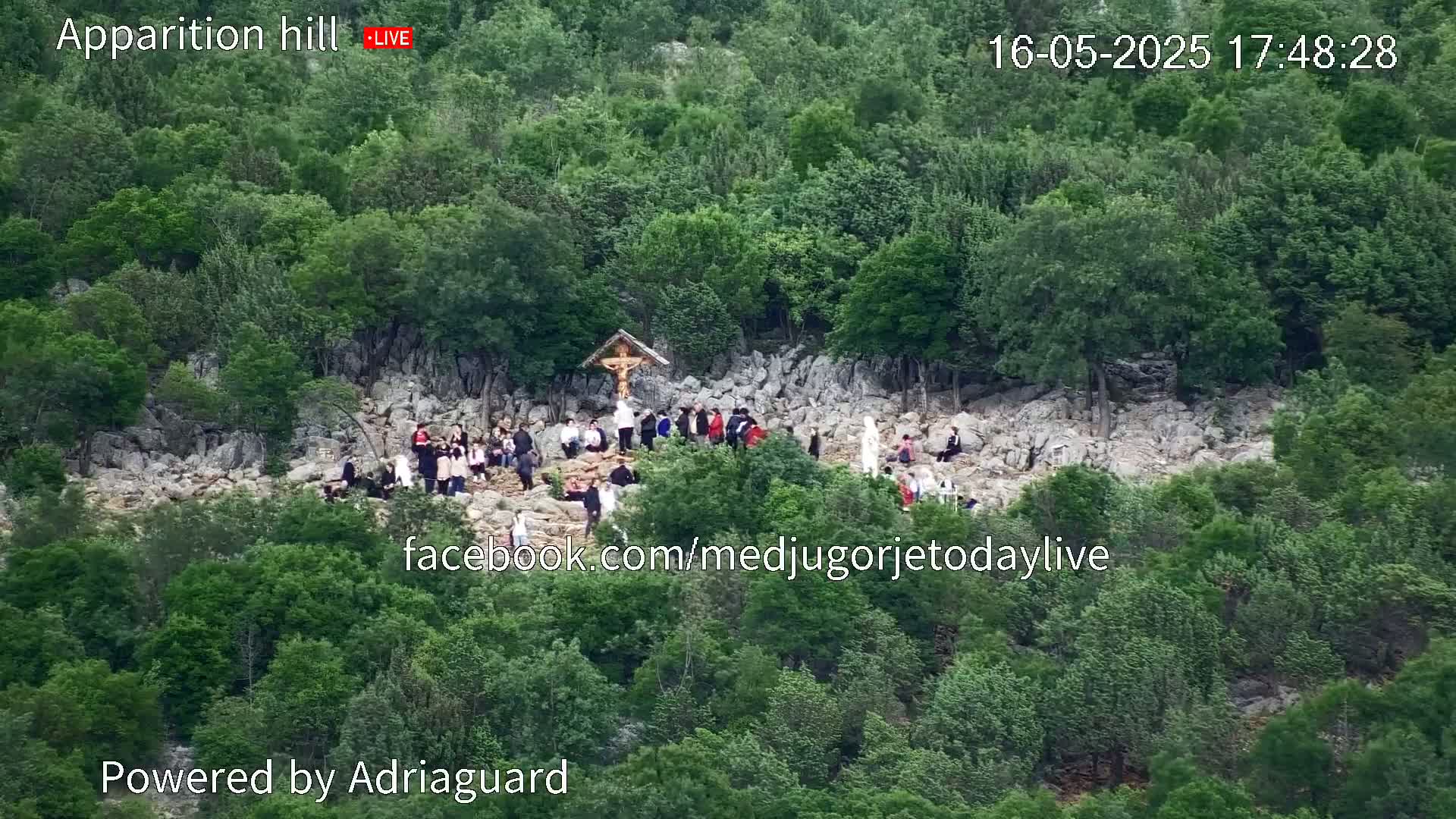 A crowd of people gathers around a crucifix on a rocky hilltop surrounded by lush green vegetation.