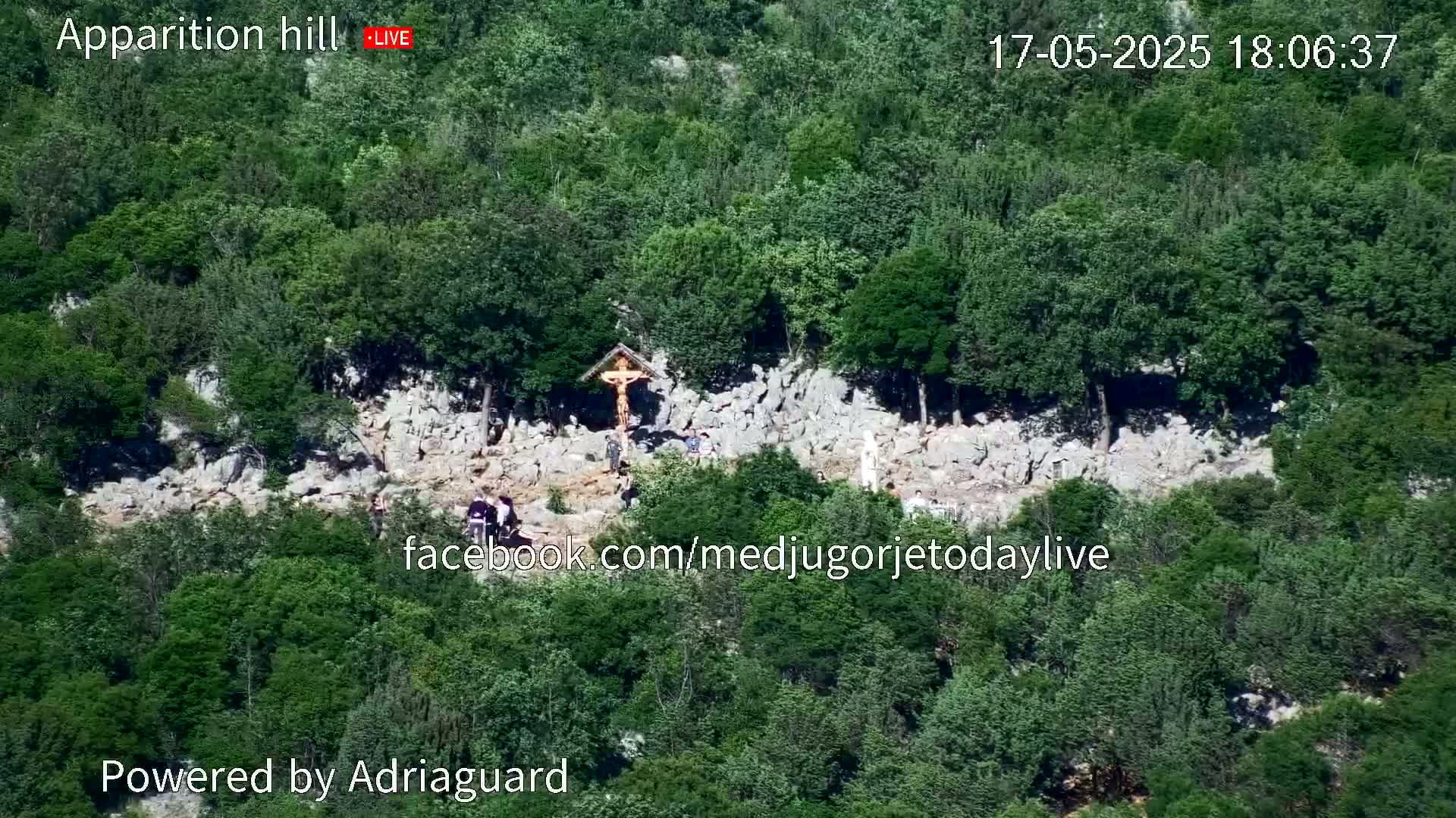 A rocky hillside covered in lush green trees and shrubs has a group of people gathered around a crucifix.