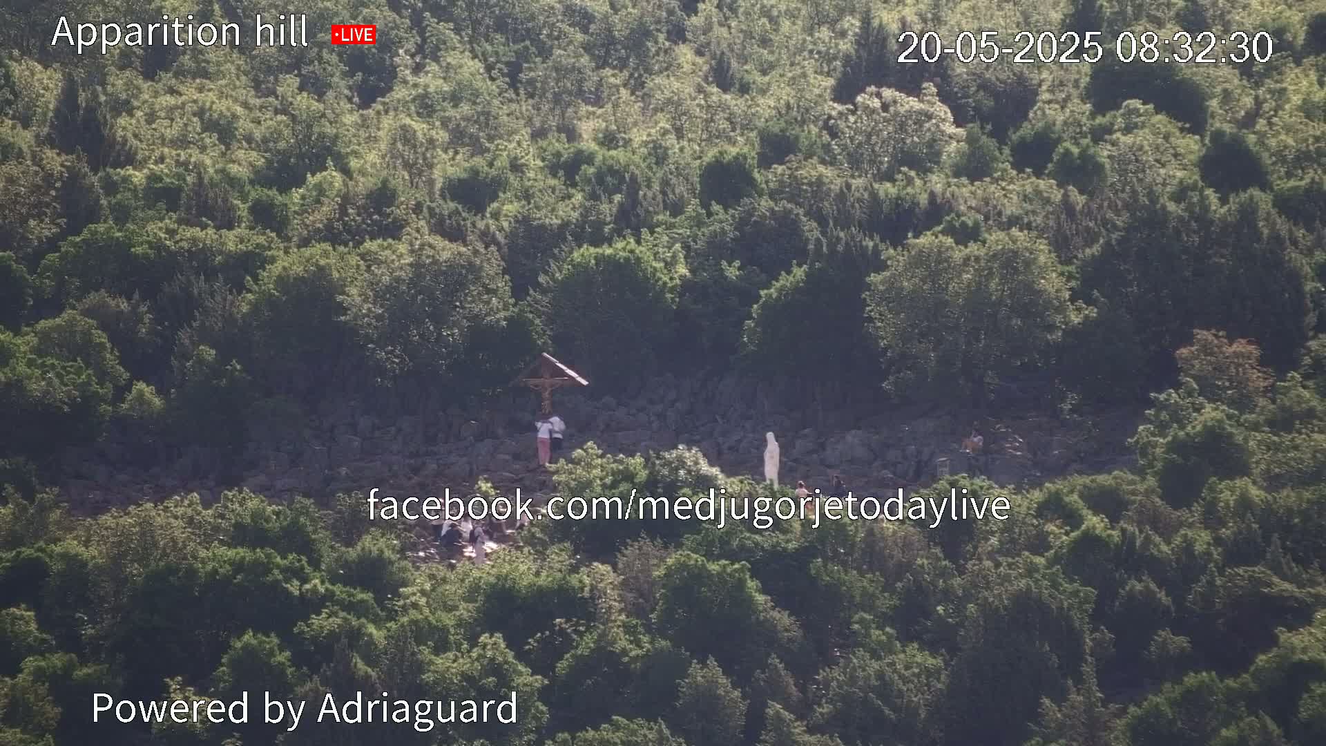 A hillside covered in green vegetation shows several people near a crucifix and statue, under a sunny sky.