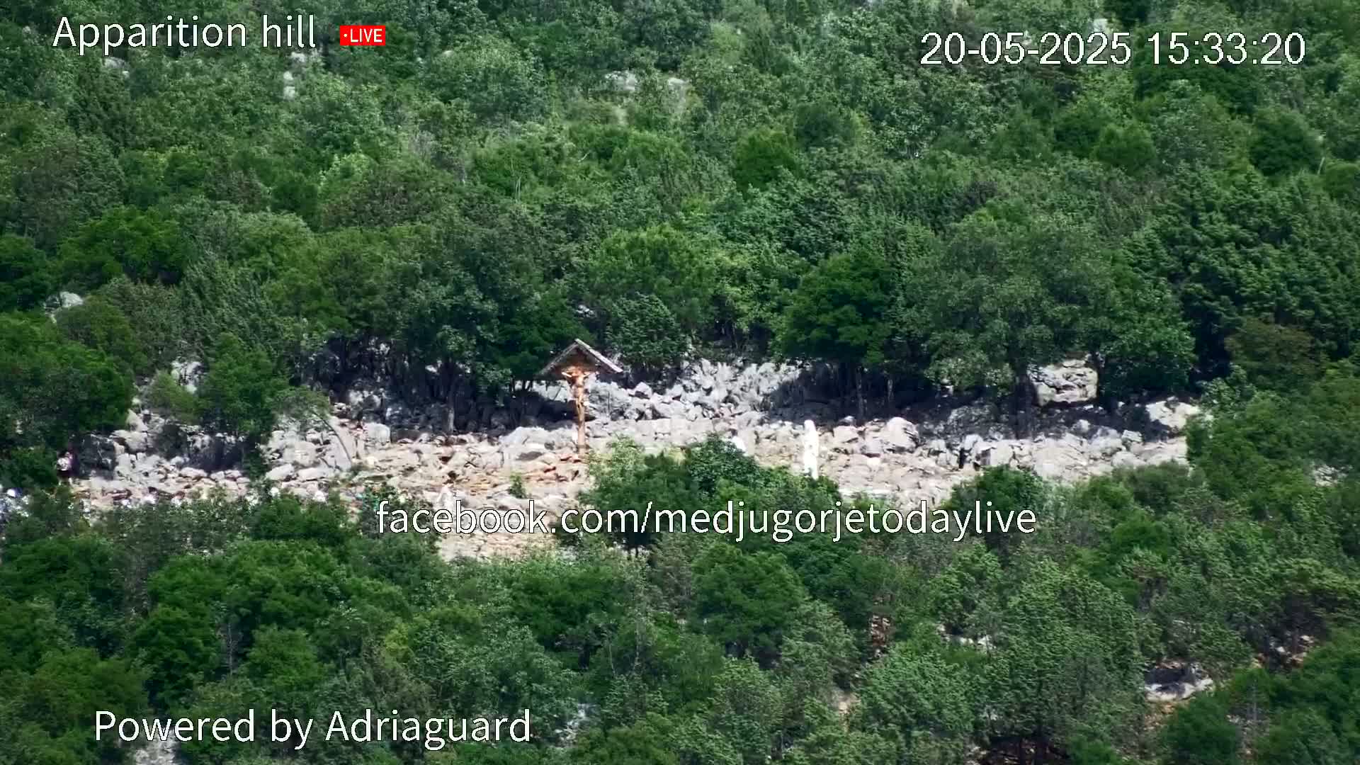 A rocky hillside covered in lush green vegetation features a crucifix and statue, under a bright, sunny sky.