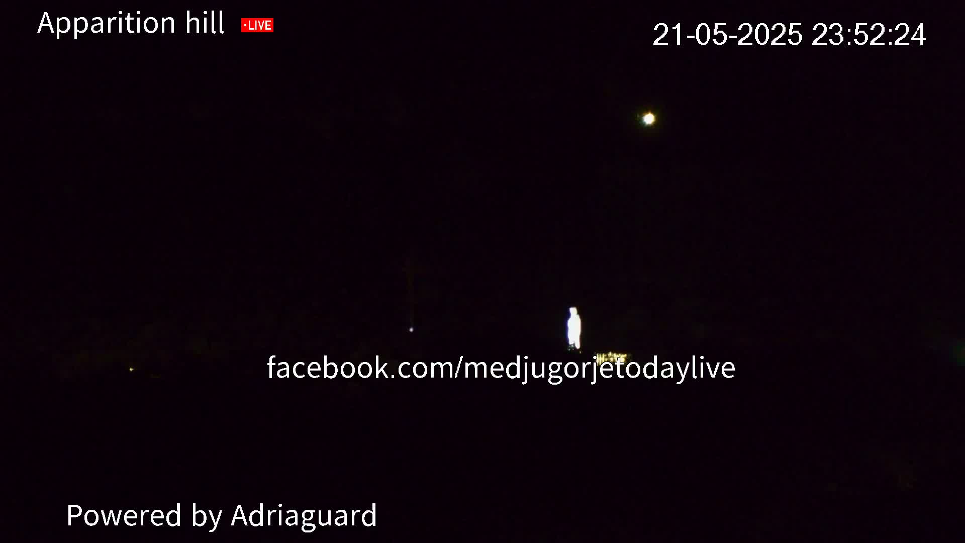 A dimly lit, nighttime scene shows a bright, human-shaped figure standing in the distance, with a bright light visible in the upper right corner of the frame.