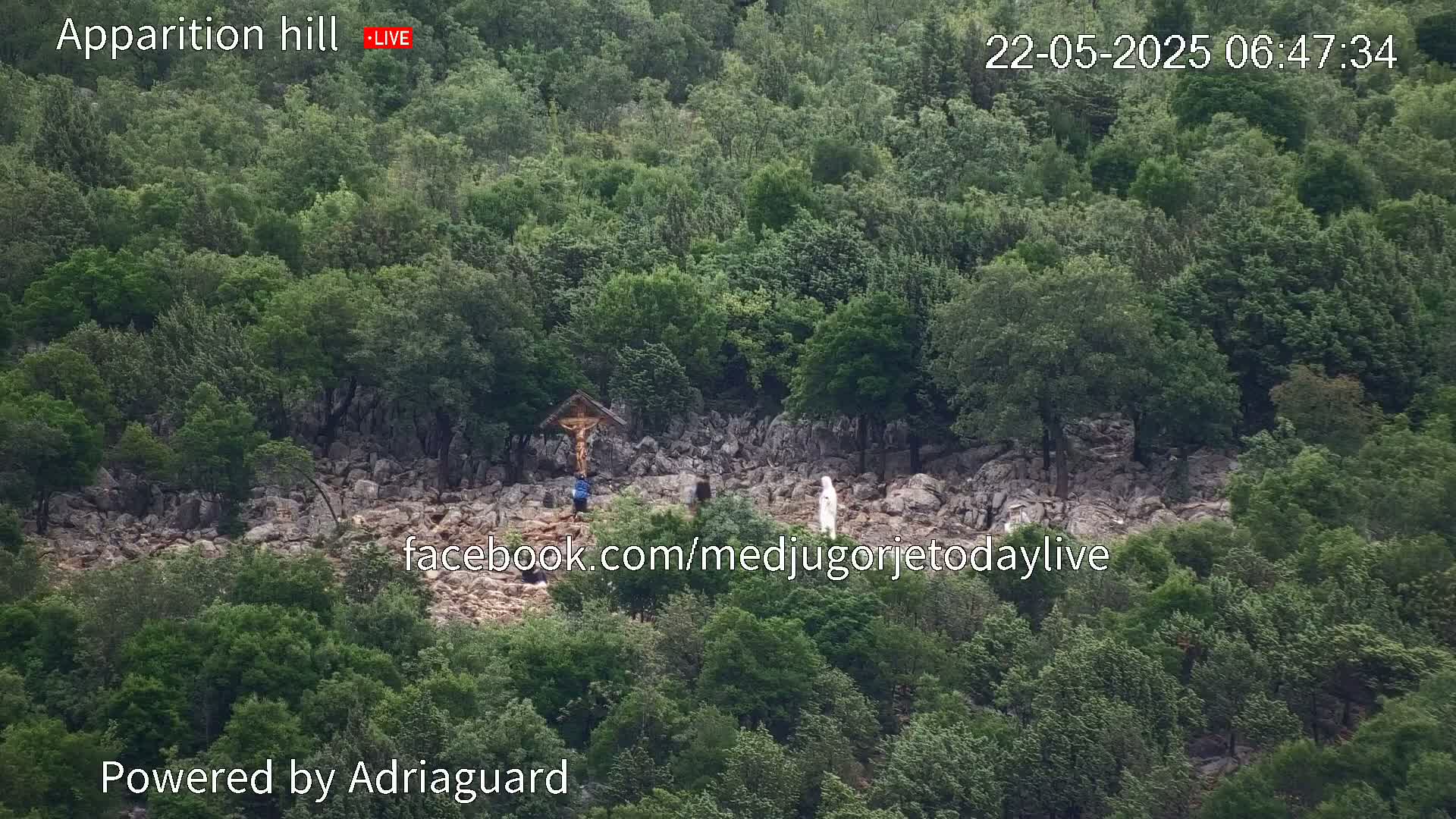 A rocky hillside covered in lush green trees, with a crucifix and a few figures visible, under an overcast sky.