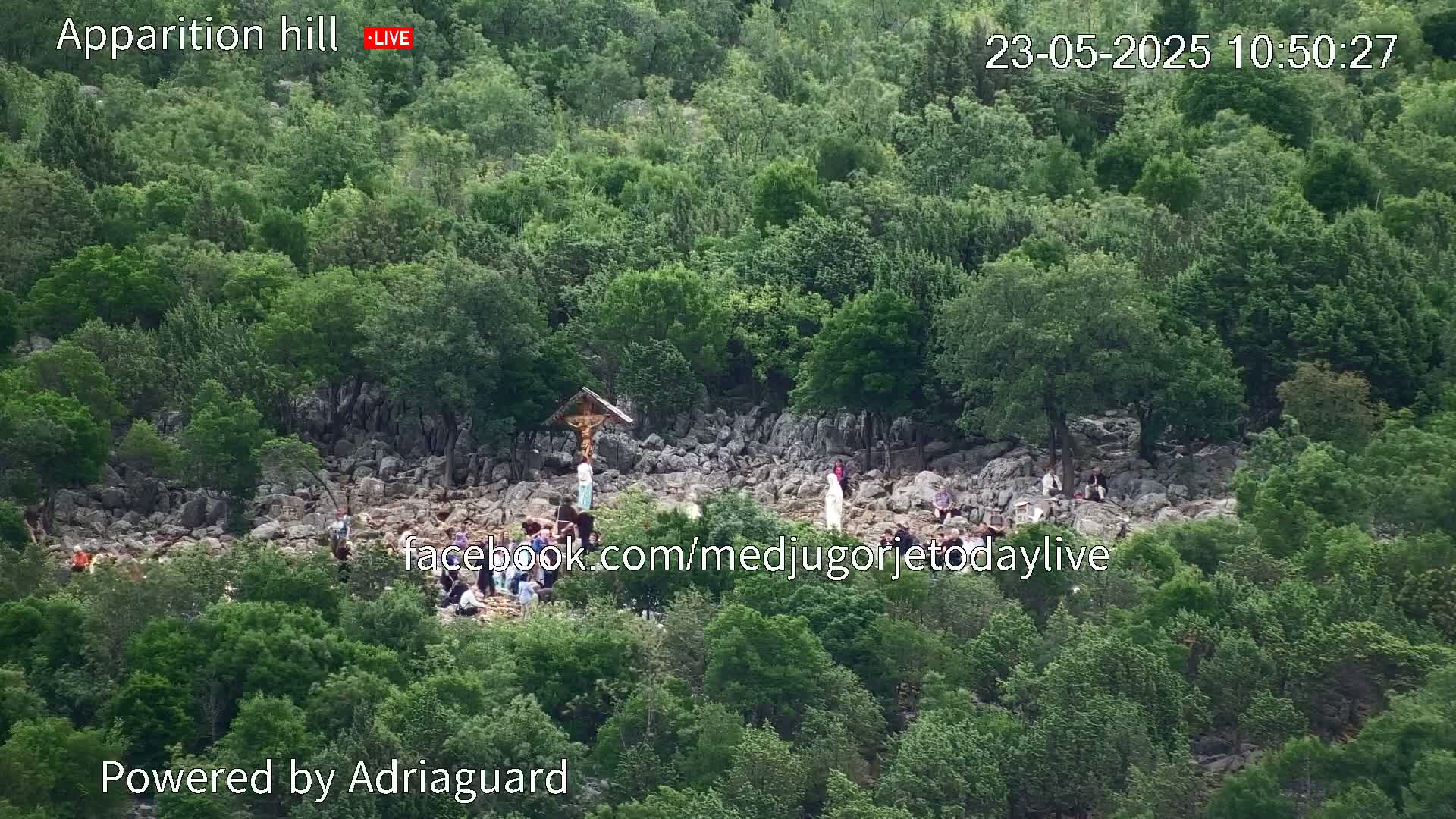 A rocky hillside covered in lush green vegetation features a crucifix and several people gathered around it, under a bright, sunny sky.