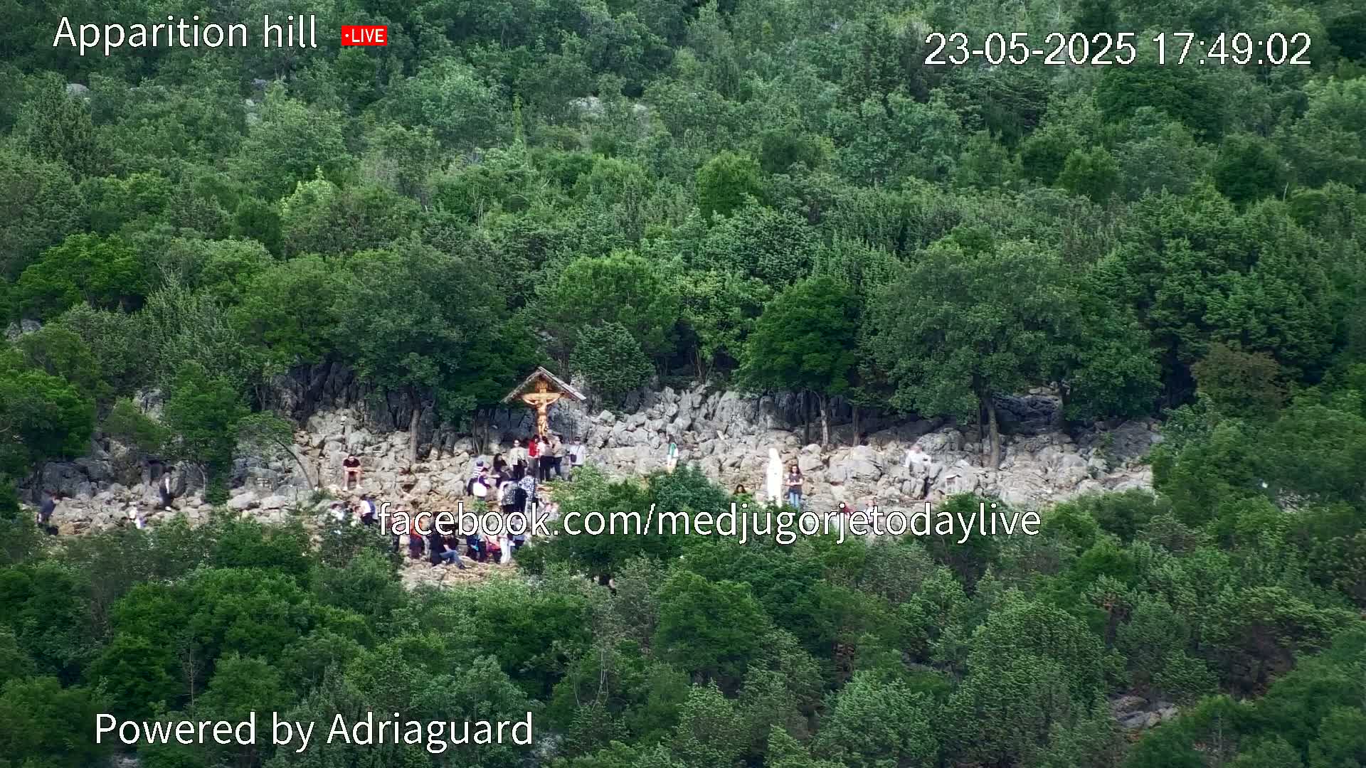 A rocky hillside covered in lush green trees, with a group of people gathered around a crucifix.