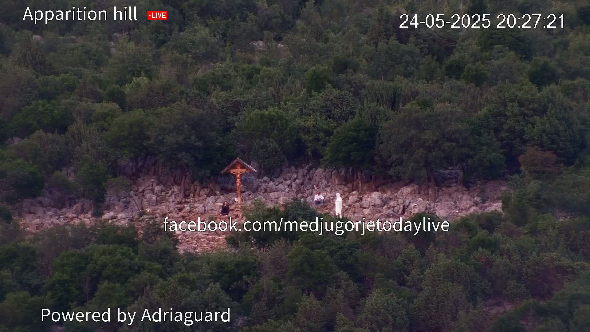 A rocky hillside covered in green vegetation shows a wooden cross and several people standing near it, under a twilight sky.