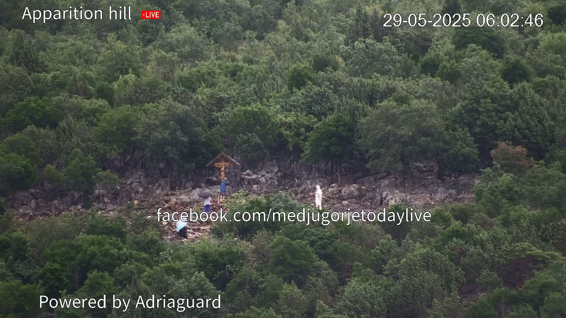 A rocky hillside covered in lush green vegetation shows several people, some under umbrellas, near a crucifix and a statue in rainy weather.