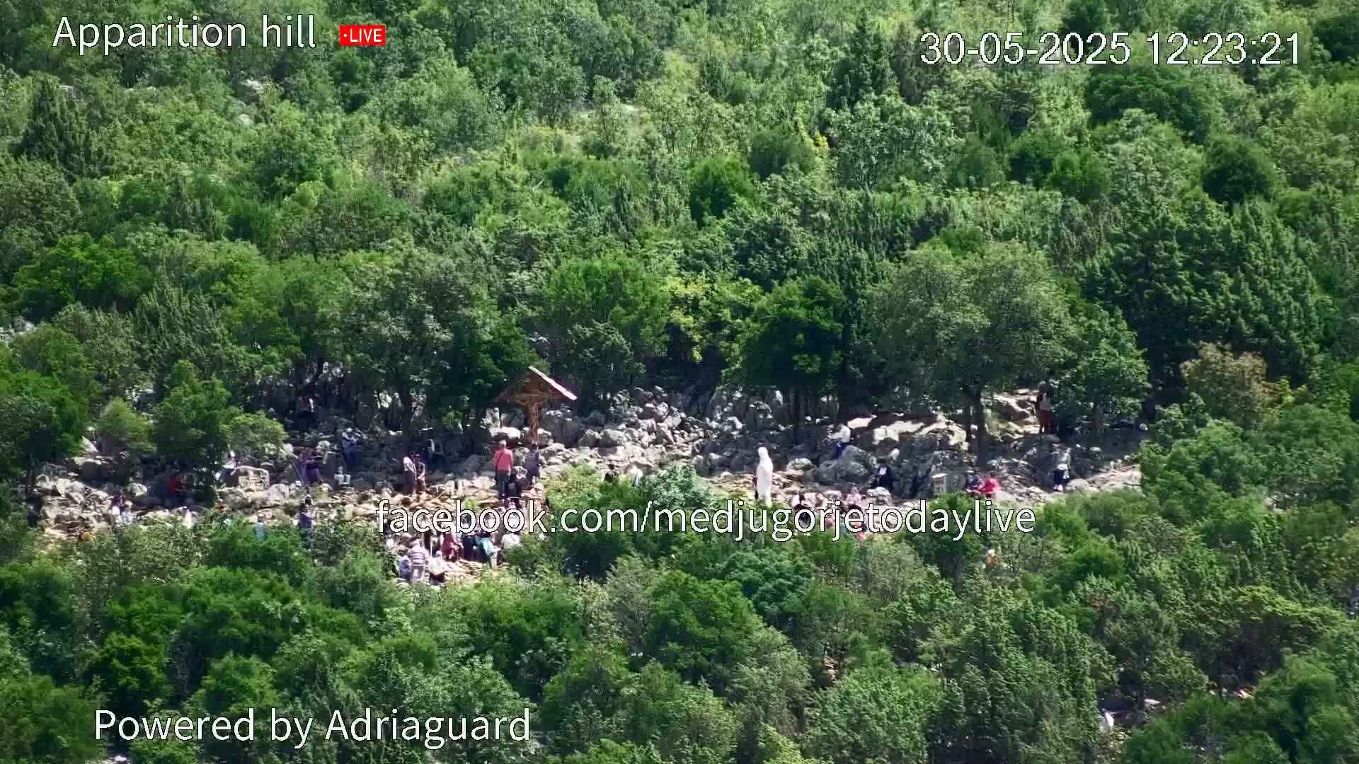 A rocky hillside covered in lush green vegetation has a number of people gathered around a small wooden structure, under a sunny sky.