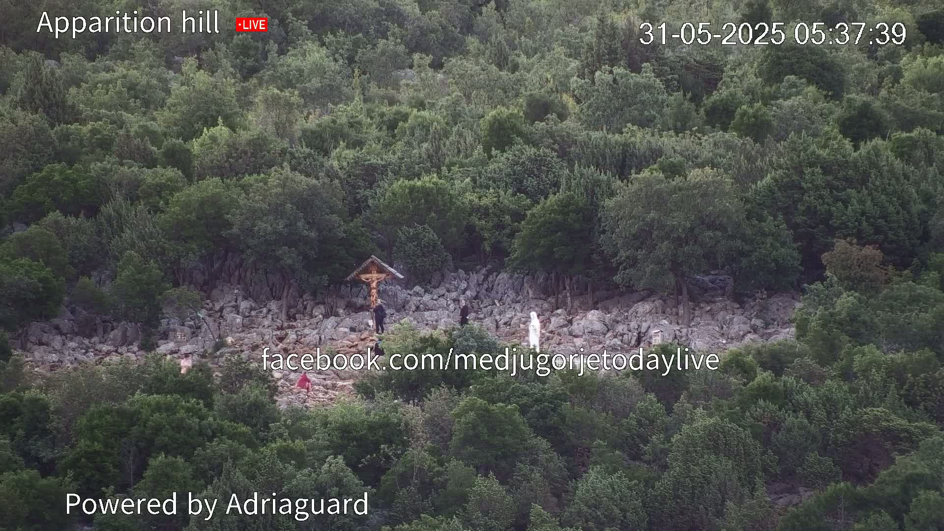 A rocky hillside covered in green trees and shrubs shows a few people near a crucifix.
