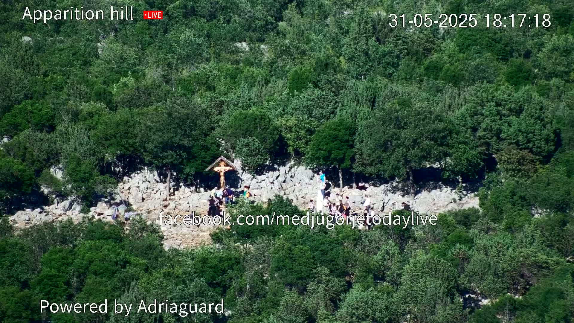 A high-angle view shows a rocky hillside covered in lush green trees, with a group of people gathered around a crucifix near the center.