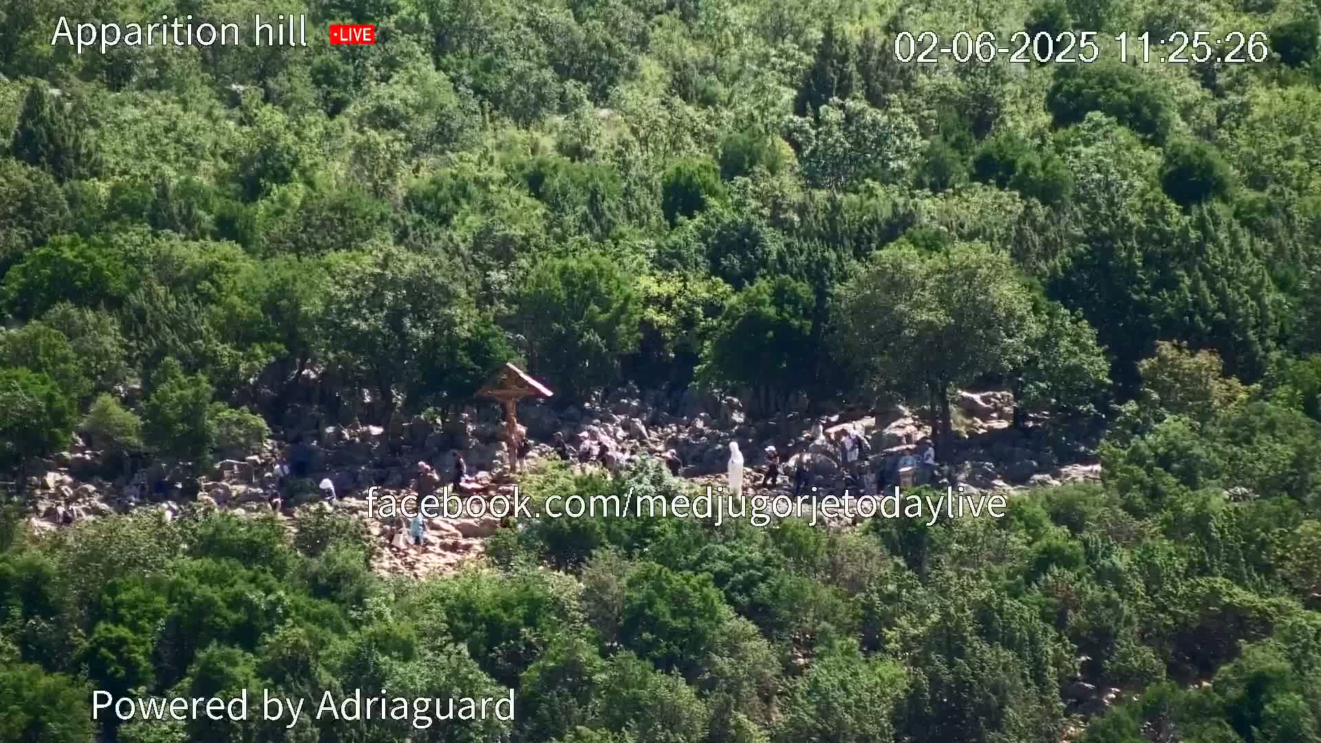 A rocky hillside covered in lush green vegetation shows a group of people gathered around a crucifix and a statue, under a bright, sunny sky.