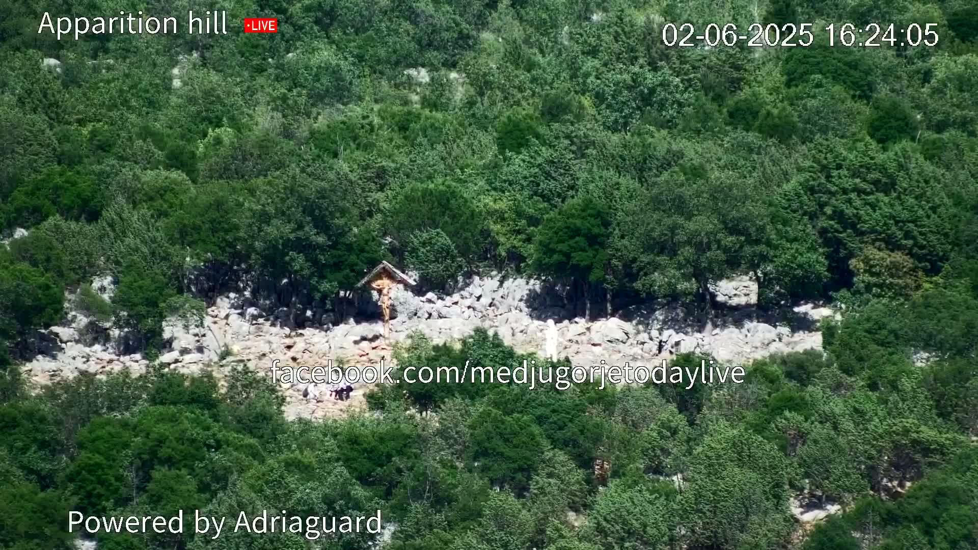 A rocky hillside covered in lush green trees and vegetation features a crucifix and small gathering of people.