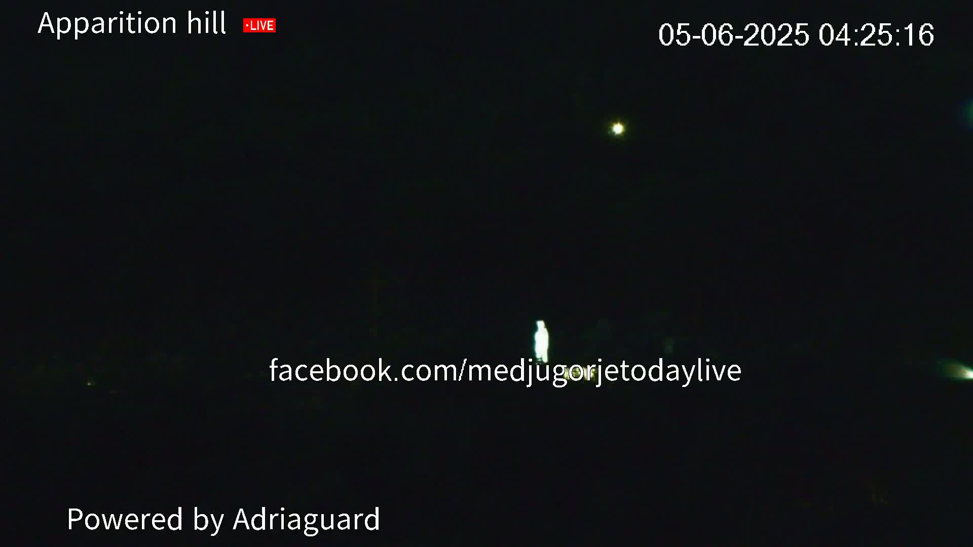 A nighttime, dark outdoor scene shows a faintly illuminated figure standing in the distance, with a few distant lights visible in the sky.