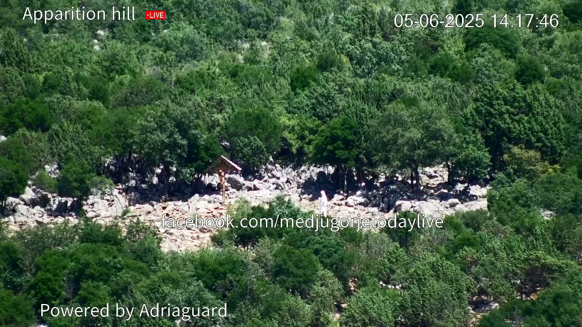 A rocky area sparsely covered with low vegetation and trees is visible, with a person in white clothing standing near a wooden cross.