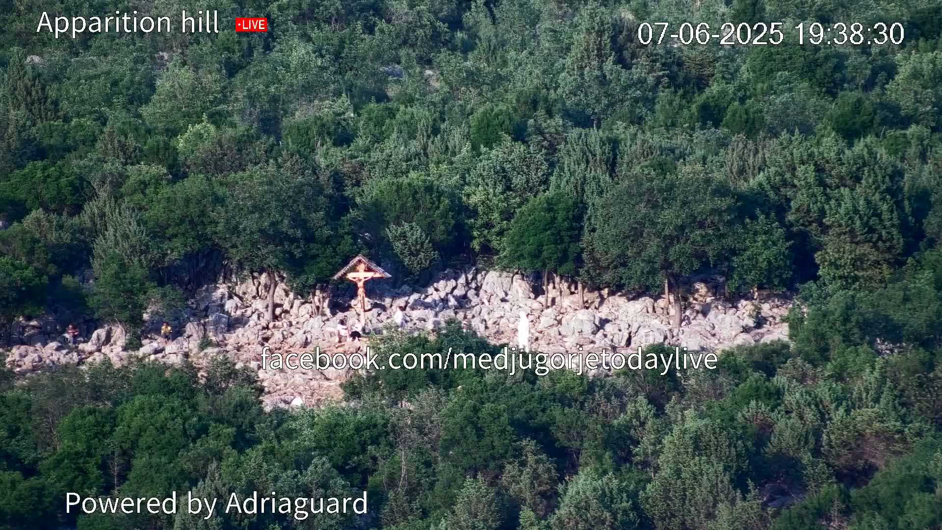 A rocky hillside covered in green trees and shrubs has a wooden crucifix and several people gathered near it.