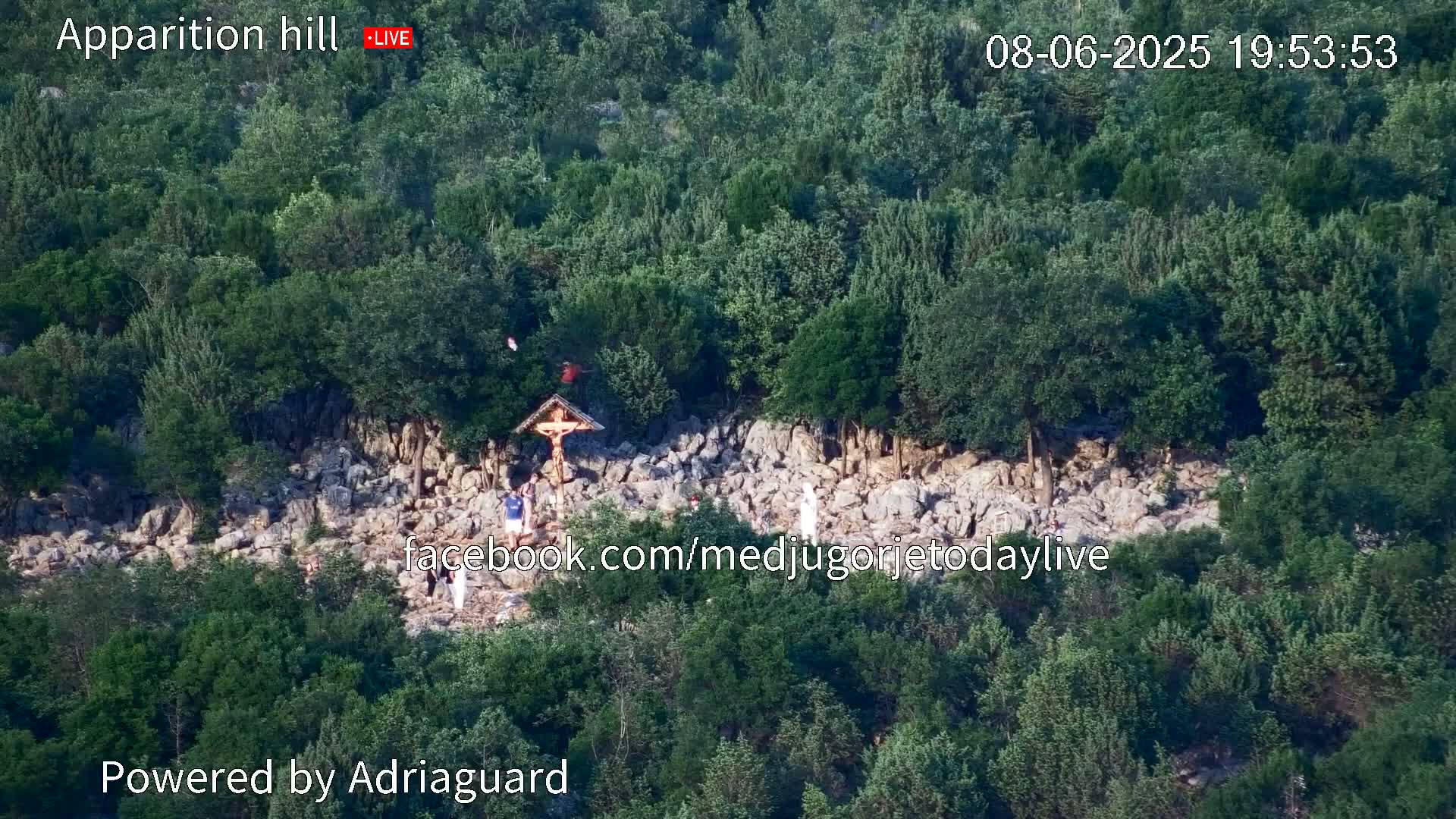 A rocky hillside covered in lush green vegetation features a crucifix and several people gathered around it.